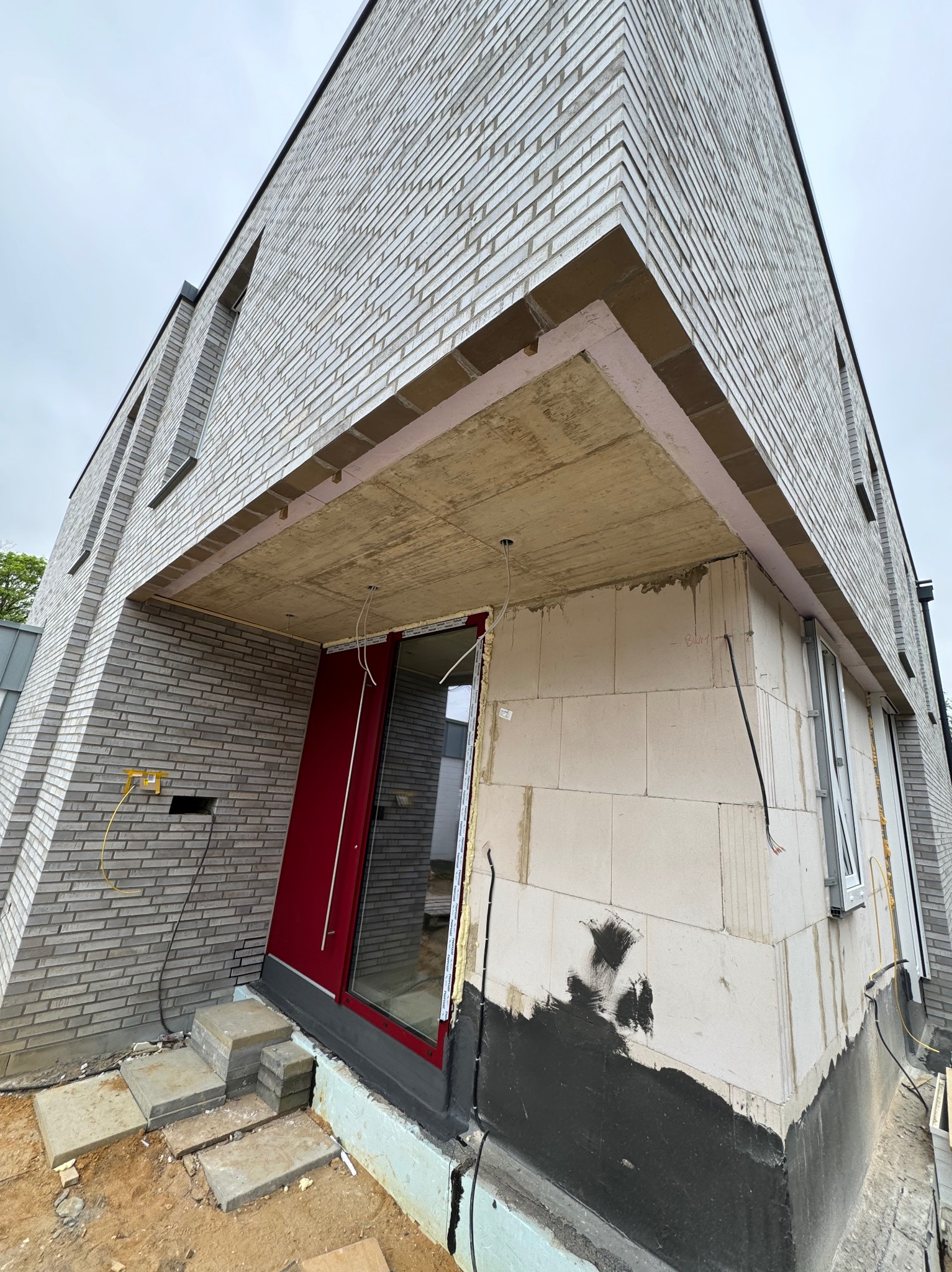 Budowa domu: surowa elewacja z szarej cegły, widoczne warstwy ocieplenia nad wejściem, czerwone drzwi wejściowe, etap prac wykończeniowych.