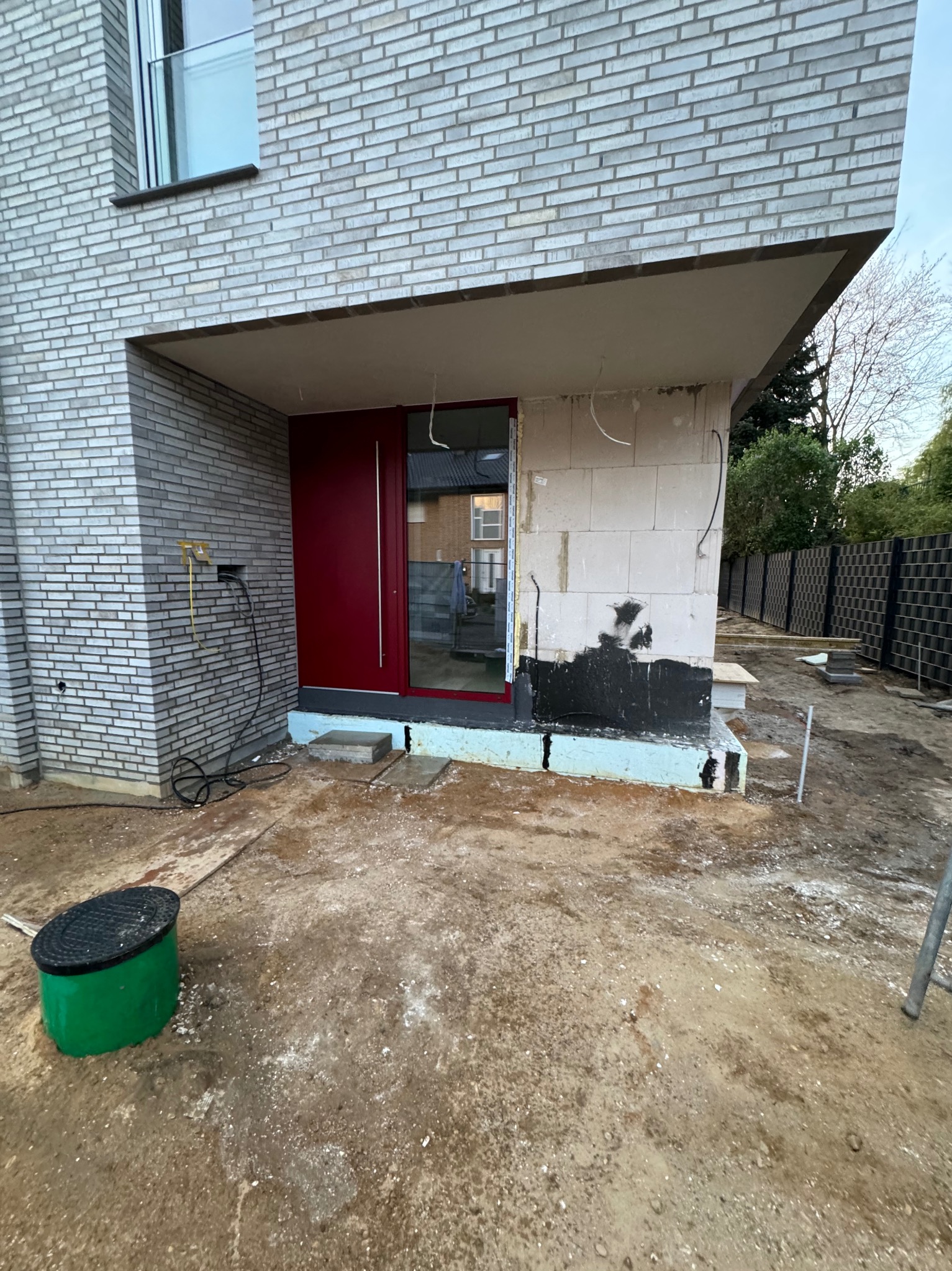 Nowoczesny dom w trakcie ocieplania elewacji: widoczna ceglana ściana, czerwone drzwi wejściowe, fragment ocieplenia przy gruncie i teren wokół domu w stanie przygotowania.