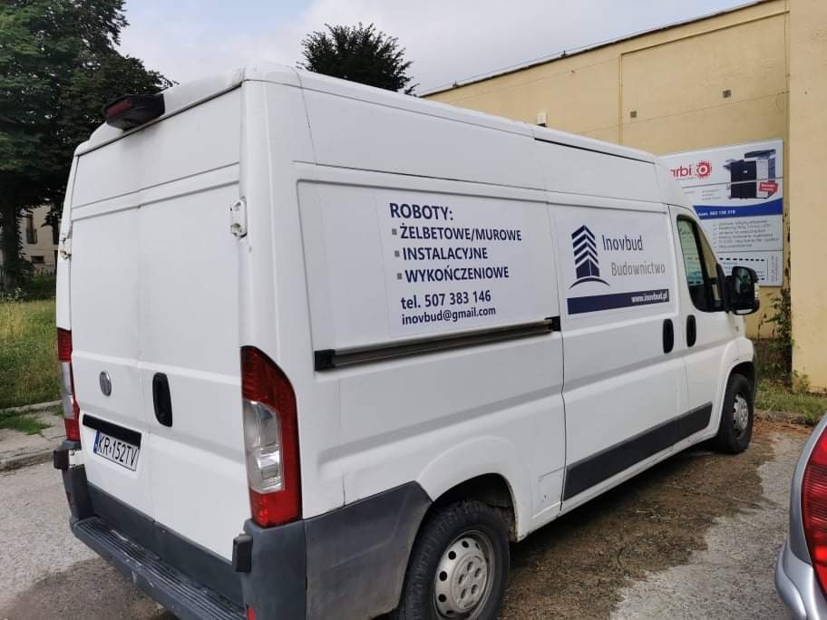 Biały samochód dostawczy firmy Inovbud Budownictwo z reklamą robót żelbetowych, murowych, instalacyjnych i wykończeniowych, zaparkowany na zewnątrz budynku.