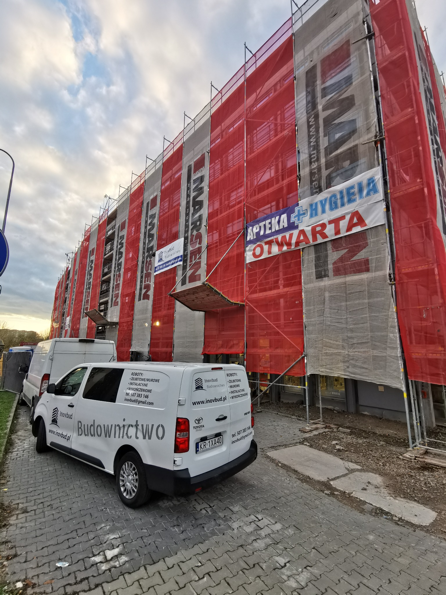 Budynek w trakcie remontu, osłonięty rusztowaniami i siatką ochronną, z zaparkowanymi samochodami dostawczymi firmy budowlanej Inovbud z widocznym logo i danymi kontaktowymi.