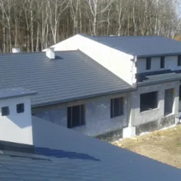 Surowy stan domu murowanego z ciemnoszarą dachówką, widok z góry na fragment dachu i komin, w tle drzewa bez liści.