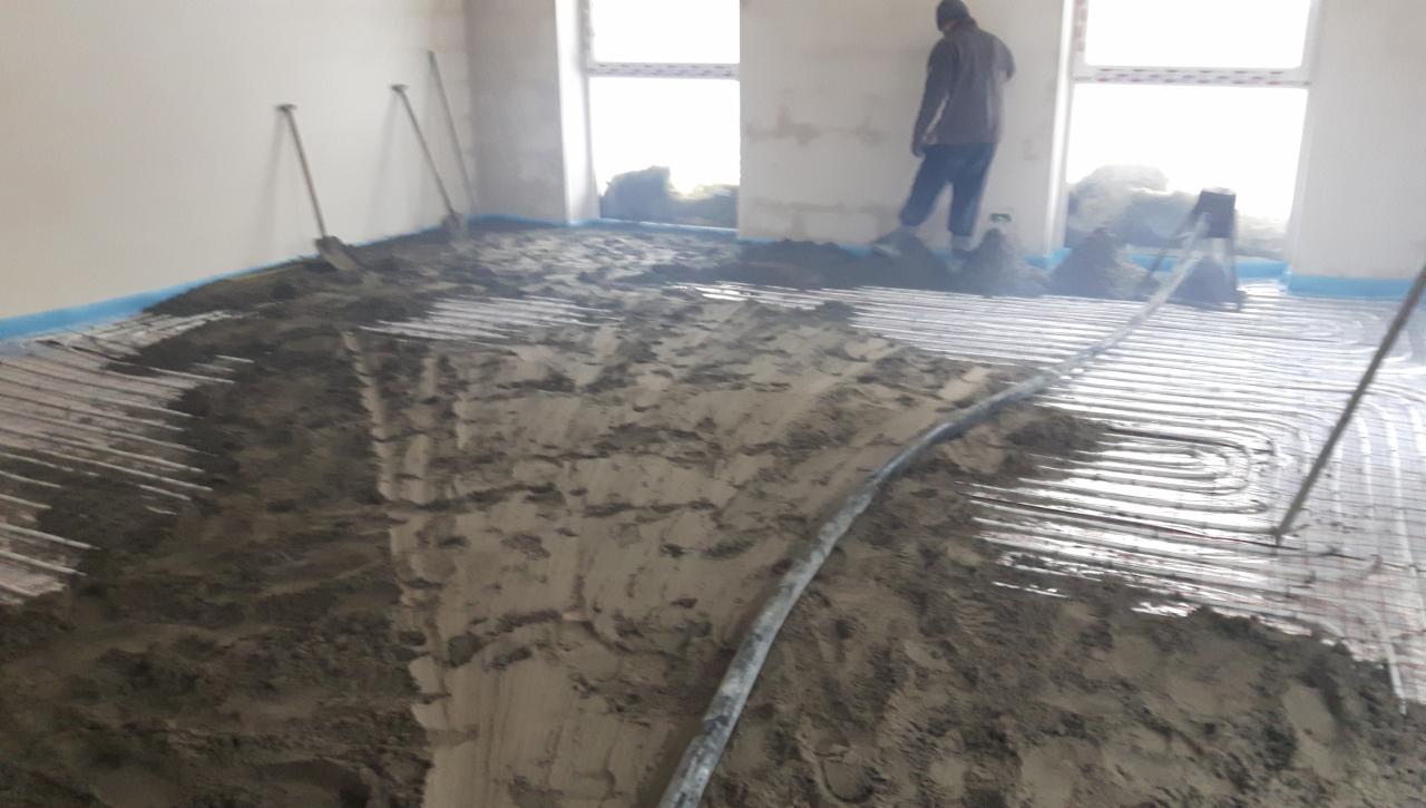 Instalacja ogrzewania podłogowego w trakcie zalewania betonem, widoczne rury grzewcze ułożone na siatce, szpachle i pracownik w tle.