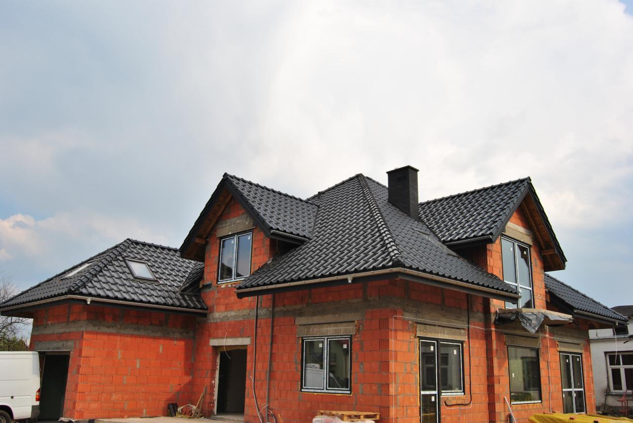 Surowy stan domu murowanego z czerwonej cegły z czarnym dachem z dachówki ceramicznej, widoczne okna z białymi ramami i czarny komin, na pierwszym planie paleta z materiałami budowlanymi, w tle...