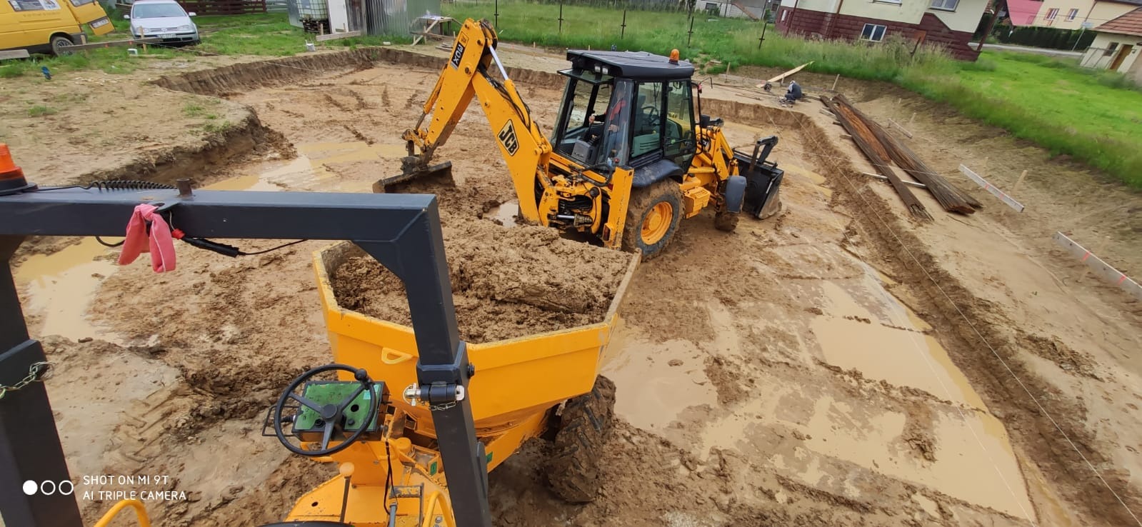 Żółta koparko-ładowarka nabiera ziemię do wywrotki na budowie, w tle widoczne przygotowane zbrojenia i wykop pod fundamenty zalany wodą.
