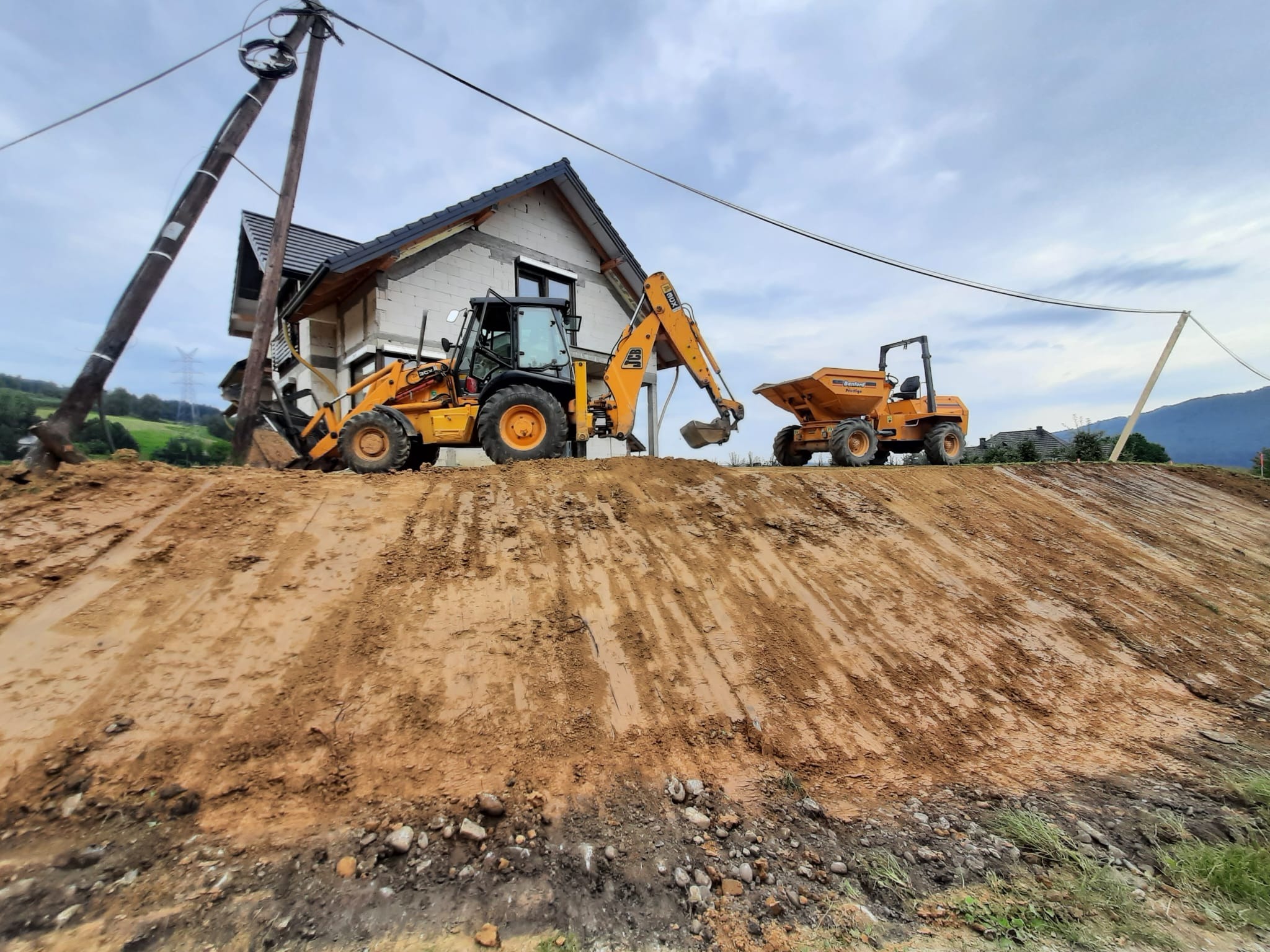 Żółta koparko-ładowarka i wywrotka na nierównym terenie budowy przed domem w stanie surowym otwartym, z pochylonym słupem energetycznym w tle.