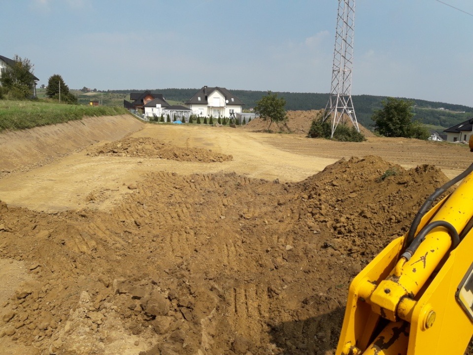 Żółty wysięgnik koparki widoczny na pierwszym planie, w tle teren w trakcie wyrównywania w Podegrodziu, z widocznymi domami i słupem wysokiego napięcia.