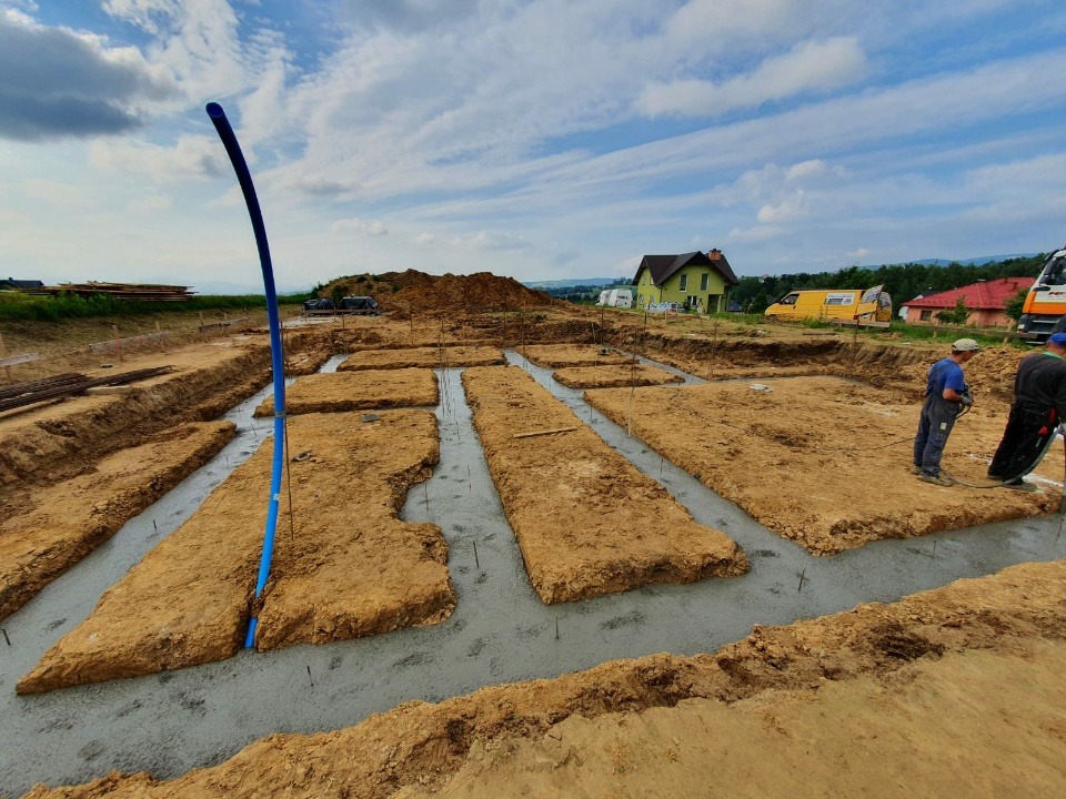 Wylane fundamenty pod budynek mieszkalny, widoczne zbrojenia i szalunki, w tle dom jednorodzinny i sprzęt budowlany.