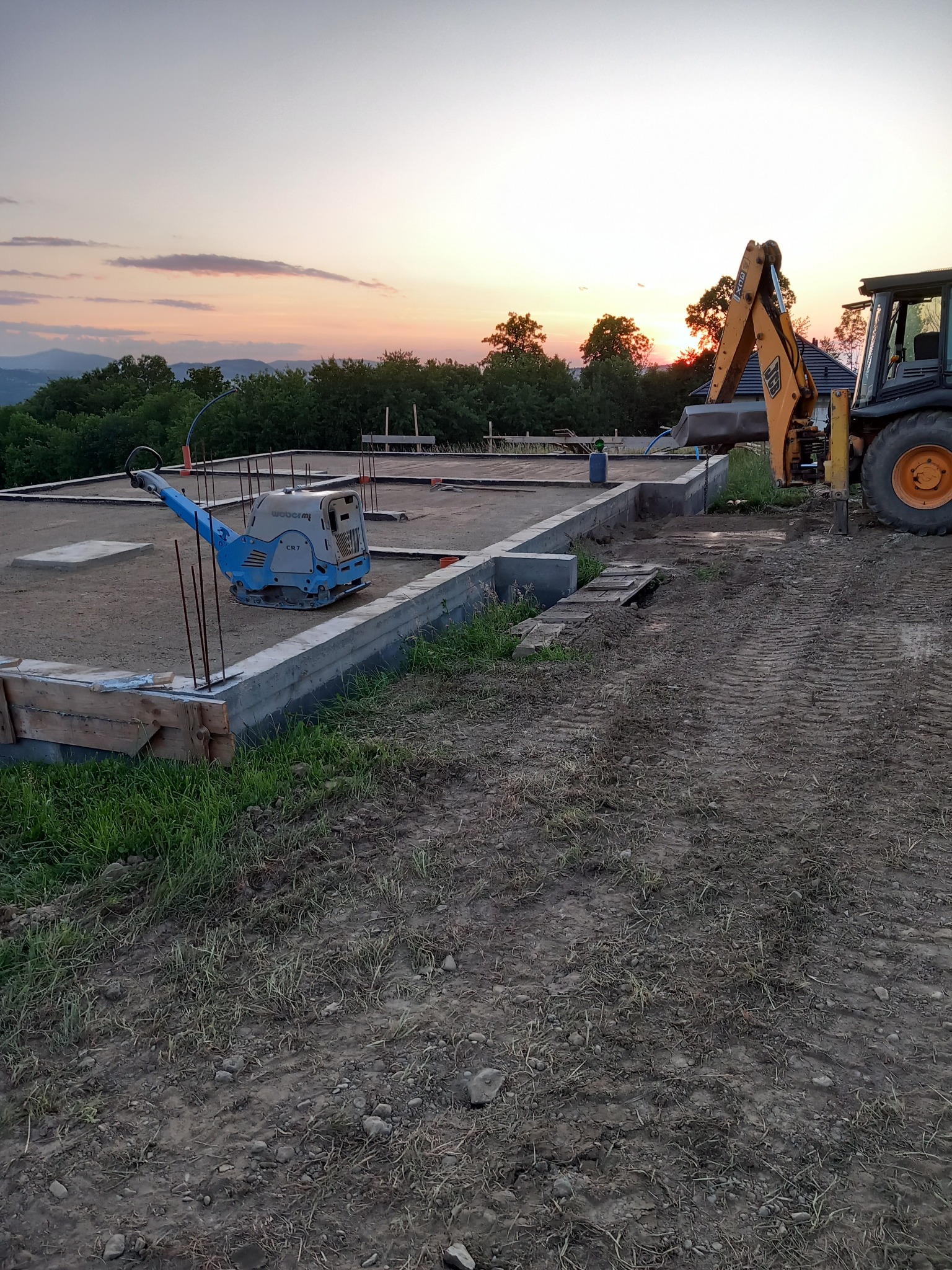 Fundamenty domu w trakcie budowy o zachodzie słońca, widoczny zagęszczarka gruntu Weber CR7 i koparko-ładowarka JCB, teren przygotowany pod wylewkę.