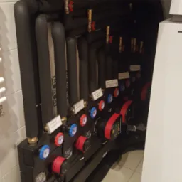 Rozdzielacz ogrzewania podłogowego z pompami obiegowymi, zaworami termostatycznymi i rurami w izolacji, zamontowany na tle białych płytek ceramicznych.