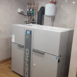 Szary piec grzewczy marki DEFRO Alpha z podłączonymi rurami i zbiornikiem wyrównawczym w pomieszczeniu wyłożonym płytkami ceramicznymi.