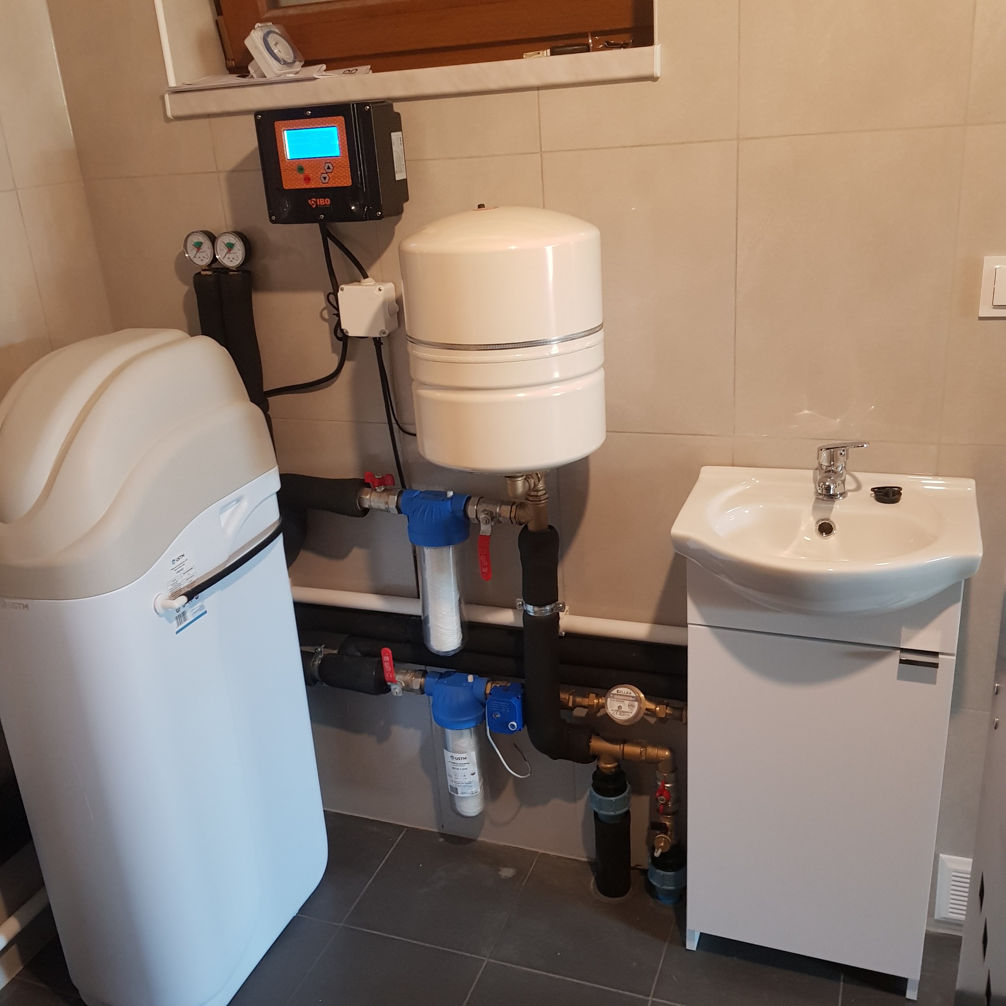 Zestaw instalacji uzdatniania wody w łazience, widoczny zmiękczacz wody, zbiornik hydroforowy, filtry mechaniczne, panel sterujący i umywalka z szafką.