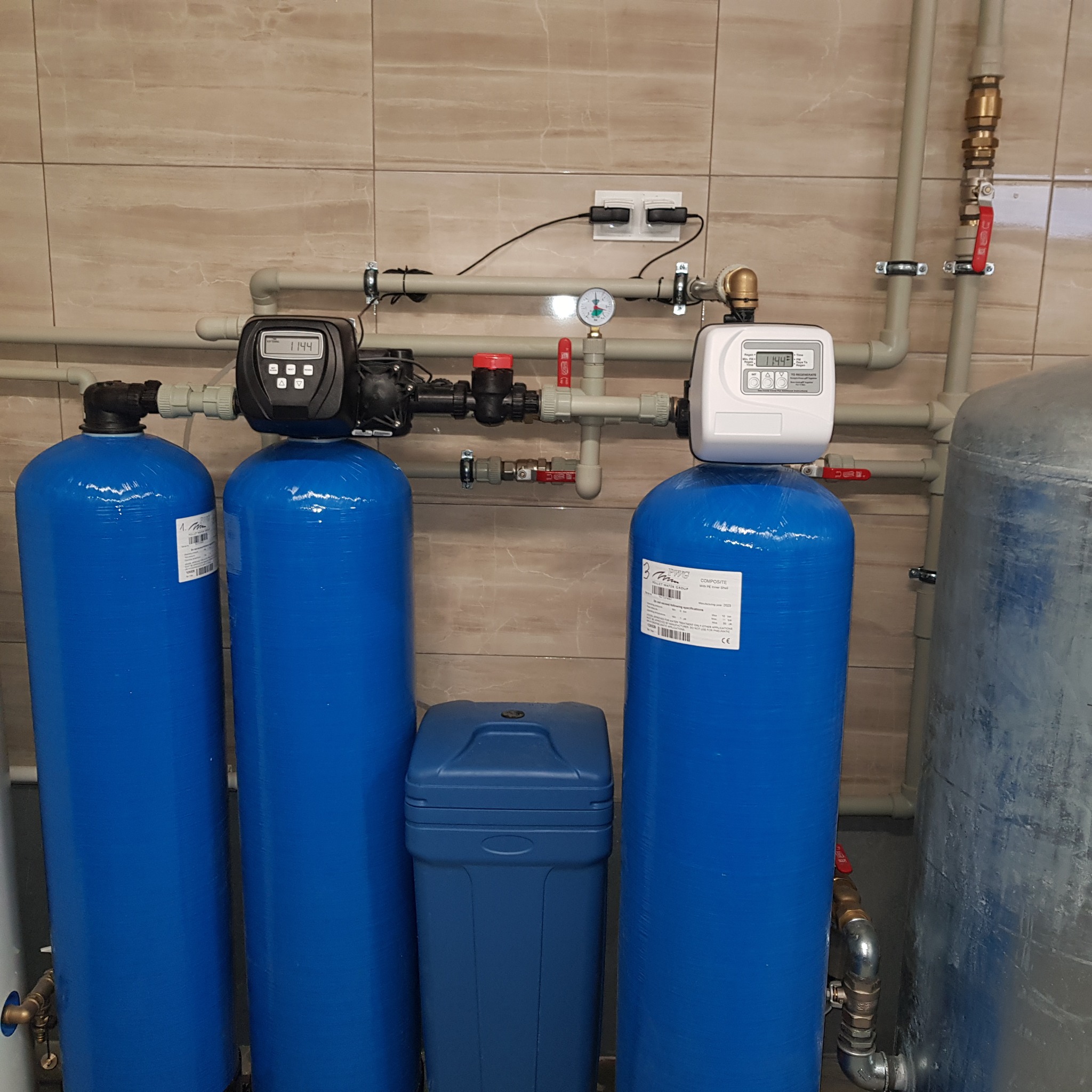 System uzdatniania wody z trzema niebieskimi zbiornikami, elektronicznymi wyświetlaczami i centralnym, niebieskim pojemnikiem na sól, zainstalowany przy beżowej ścianie z płytek.