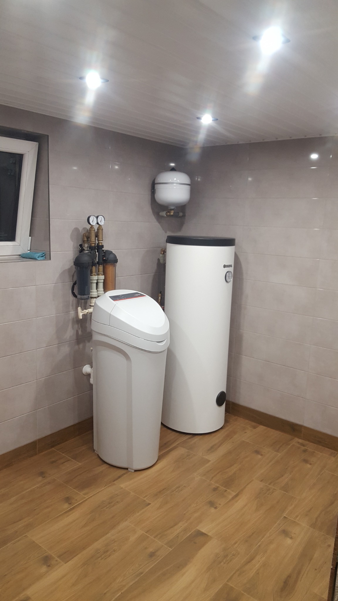 Biały zmiękczacz wody i pionowy bojler w pomieszczeniu z jasnymi płytkami na ścianach i podłodze imitującej drewno, widoczne filtry i manometry na ścianie.