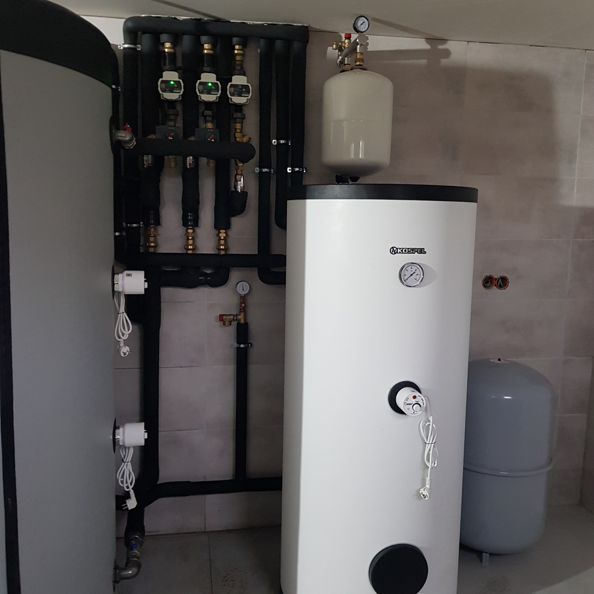 Biały, pionowy zbiornik Kospel z czarnym panelem, podłączony do czarnych rur instalacji grzewczej z zielonymi przepływomierzami, na tle płytek ceramicznych.