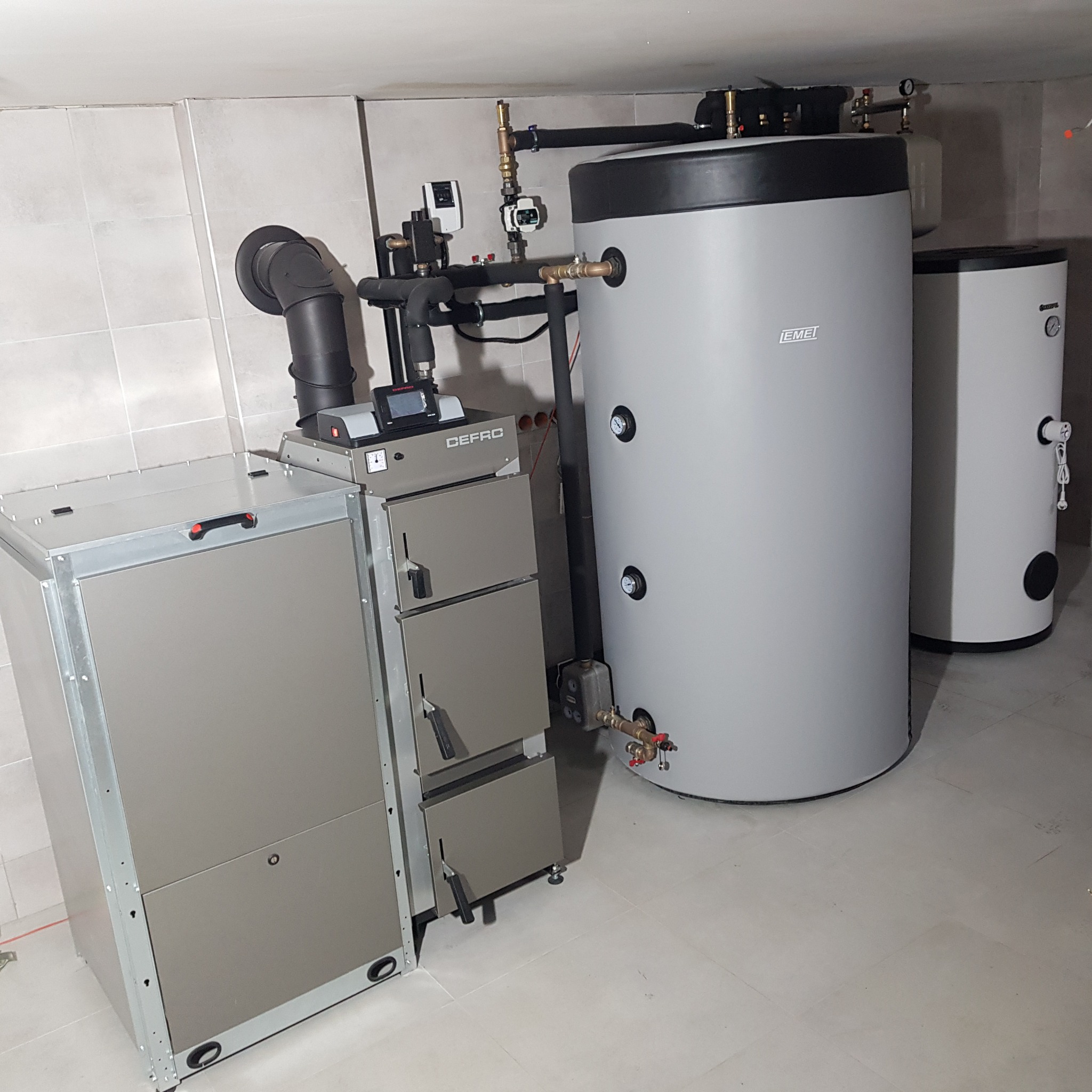 Nowoczesna kotłownia z kotłem na paliwo stałe marki DEFRO, zbiornikiem buforowym EMET i dodatkowym zasobnikiem ciepłej wody w pomieszczeniu wyłożonym płytkami ceramicznymi.