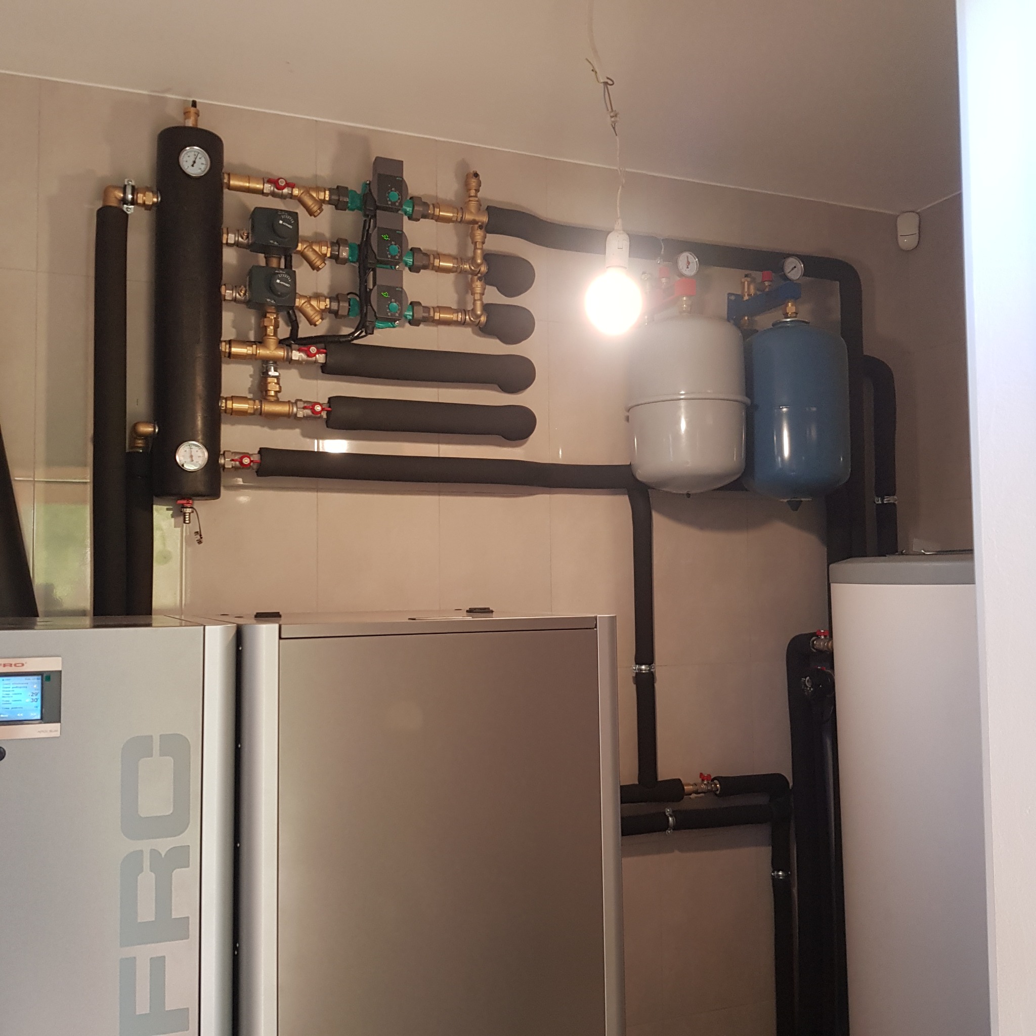 Węzeł cieplny z czarnymi rurami izolowanymi, manometrami, zaworami i dwoma zbiornikami wyrównawczymi w kolorach szarym i niebieskim, podwieszona żarówka w tle.