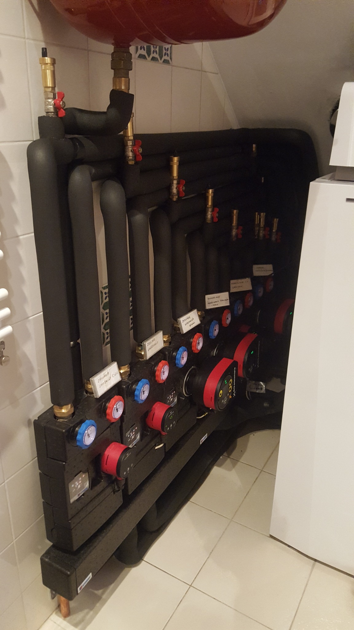 Rozdzielacz ogrzewania podłogowego z pompami obiegowymi, zaworami termostatycznymi i rurami w izolacji, zamontowany na tle białych płytek ceramicznych.