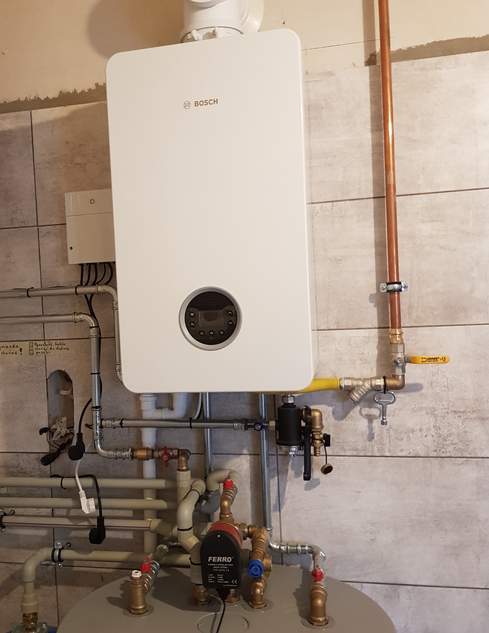 Biały, wiszący piec gazowy marki Bosch z podłączeniami hydraulicznymi i elektrycznymi, widoczne rury miedziane i plastikowe, pompa obiegowa Ferro, zamontowany na ścianie z szarych płytek.