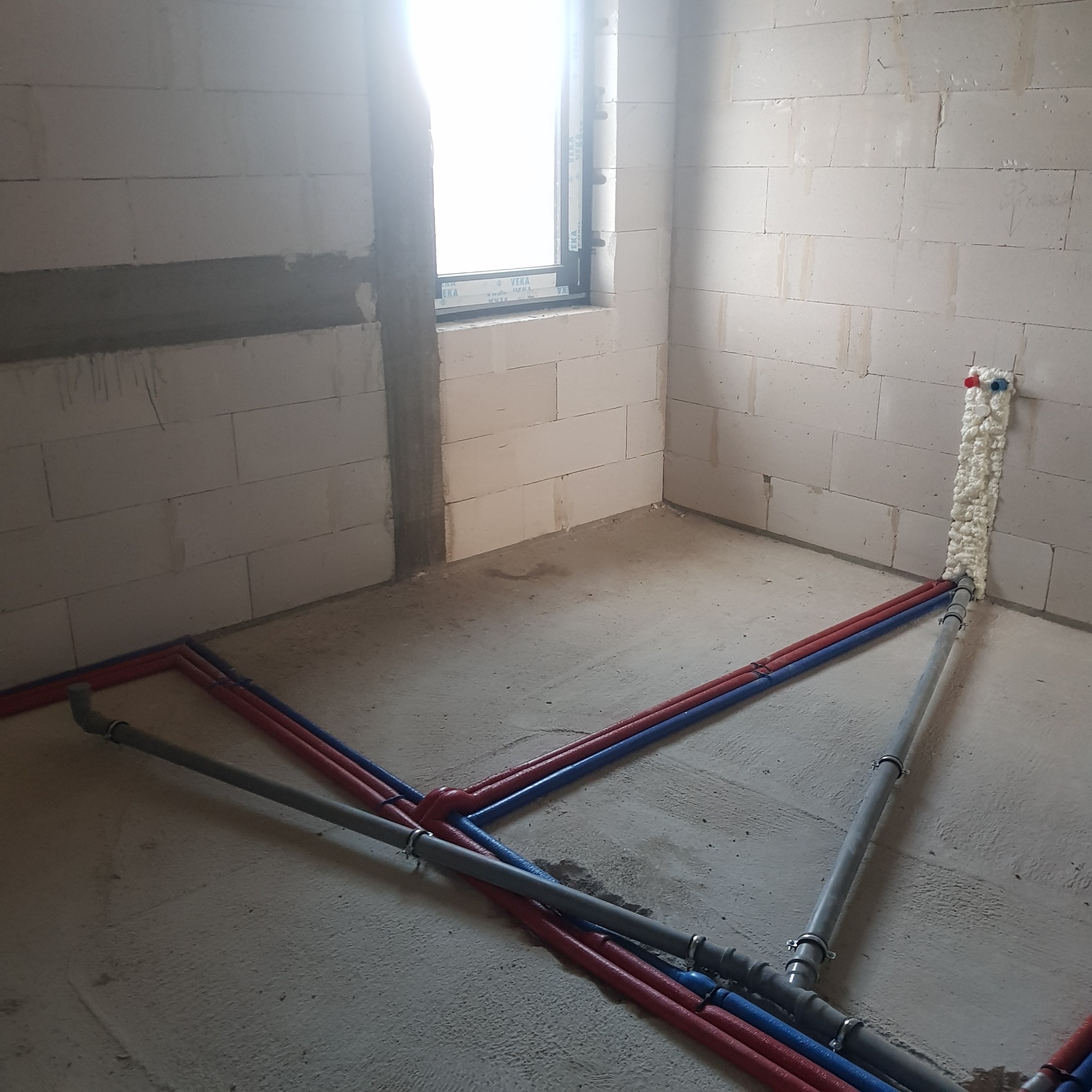 Surowy stan instalacji wodnej i kanalizacyjnej w nowym budynku: czerwone i niebieskie rury wodne oraz szare rury kanalizacyjne prowadzone po podłodze w kierunku ściany, zabezpieczone pianką...