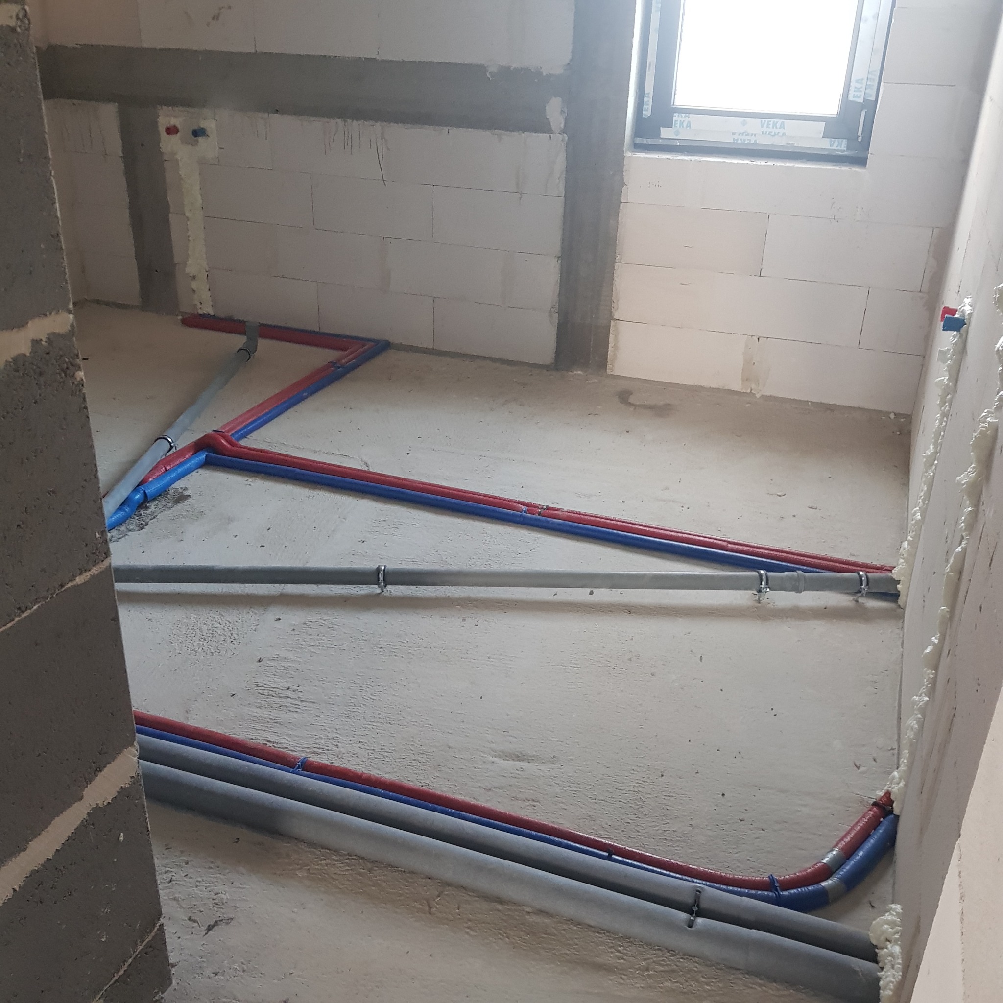 Wnętrze w trakcie prac hydraulicznych: rury wodne (czerwone i niebieskie) oraz szare rury kanalizacyjne ułożone na surowej posadzce, pianka montażowa uszczelniająca przejścia przez ściany z betonu...