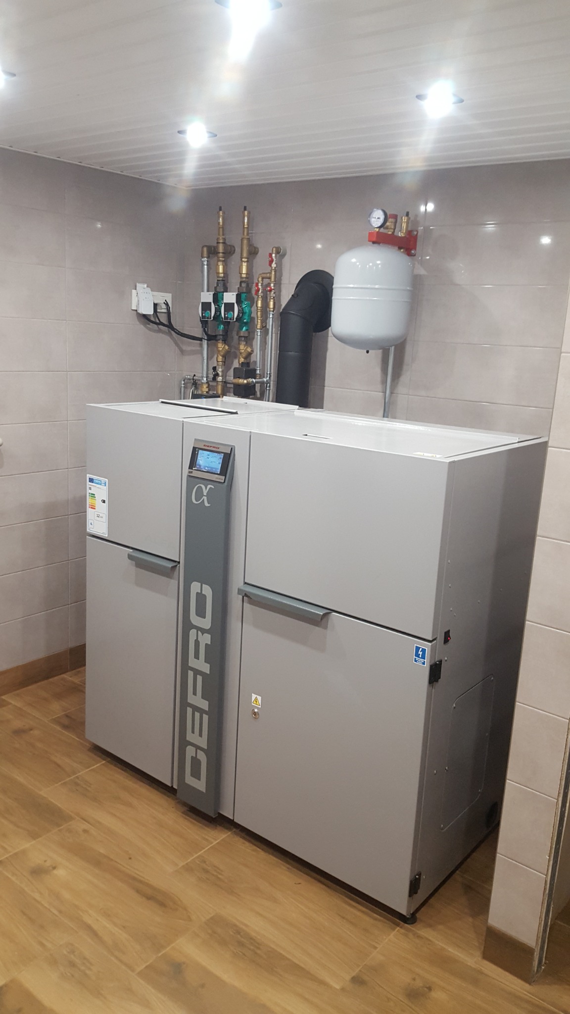 Szary piec grzewczy marki DEFRO Alpha z podłączonymi rurami i zbiornikiem wyrównawczym w pomieszczeniu wyłożonym płytkami ceramicznymi.