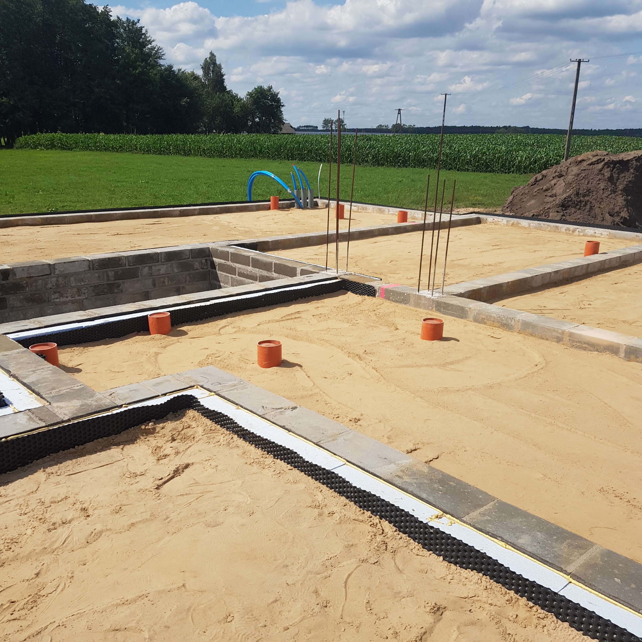 Fundamenty domu jednorodzinnego w trakcie budowy, z widoczną izolacją pionową, szalunkami i elementami instalacji. Tło stanowi zielone pole i błękitne niebo.