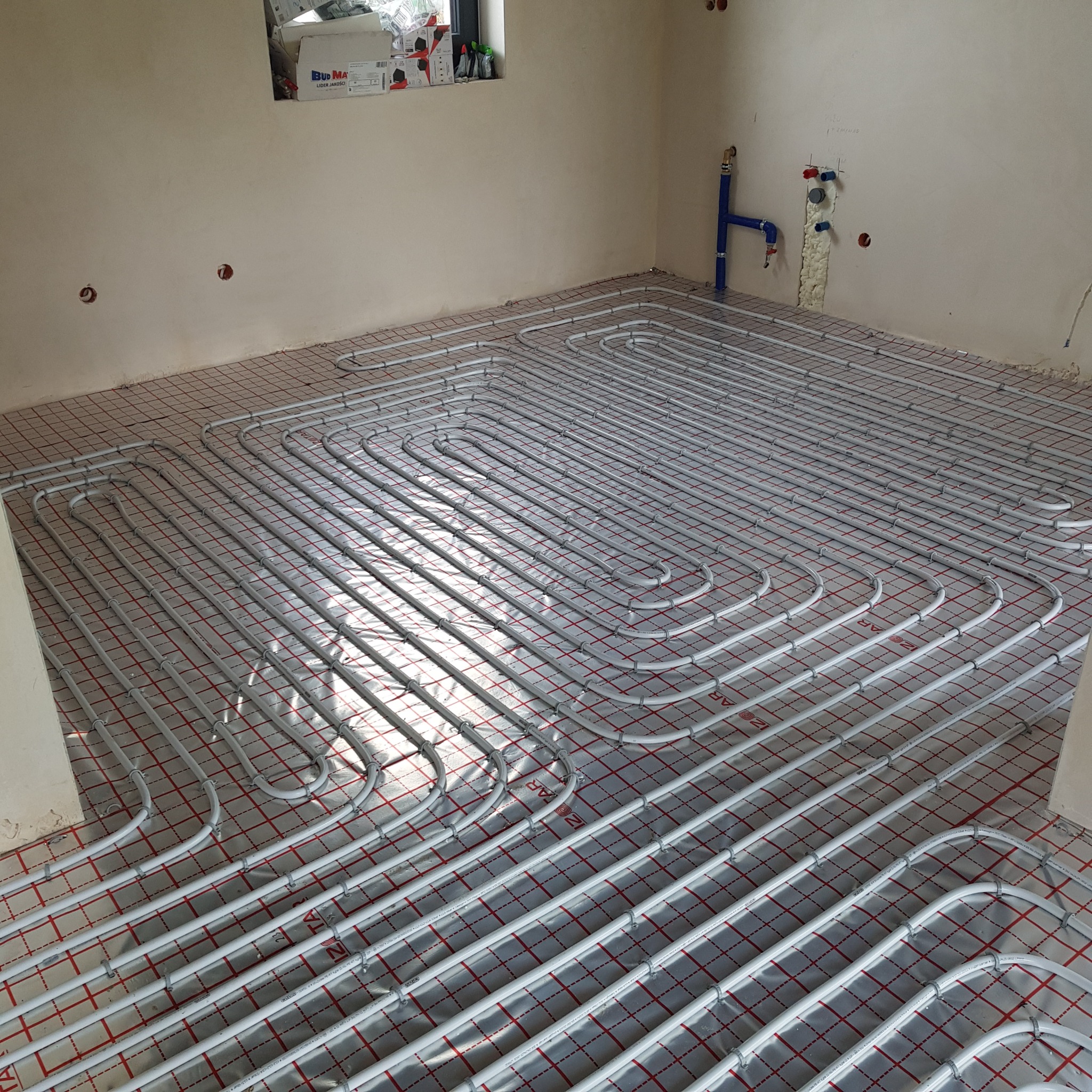 Ułożone rury ogrzewania podłogowego na folii z siatką w surowym wnętrzu. Widoczne niebieskie rury instalacji wodnej przy ścianie. Etap przed wylewką.
