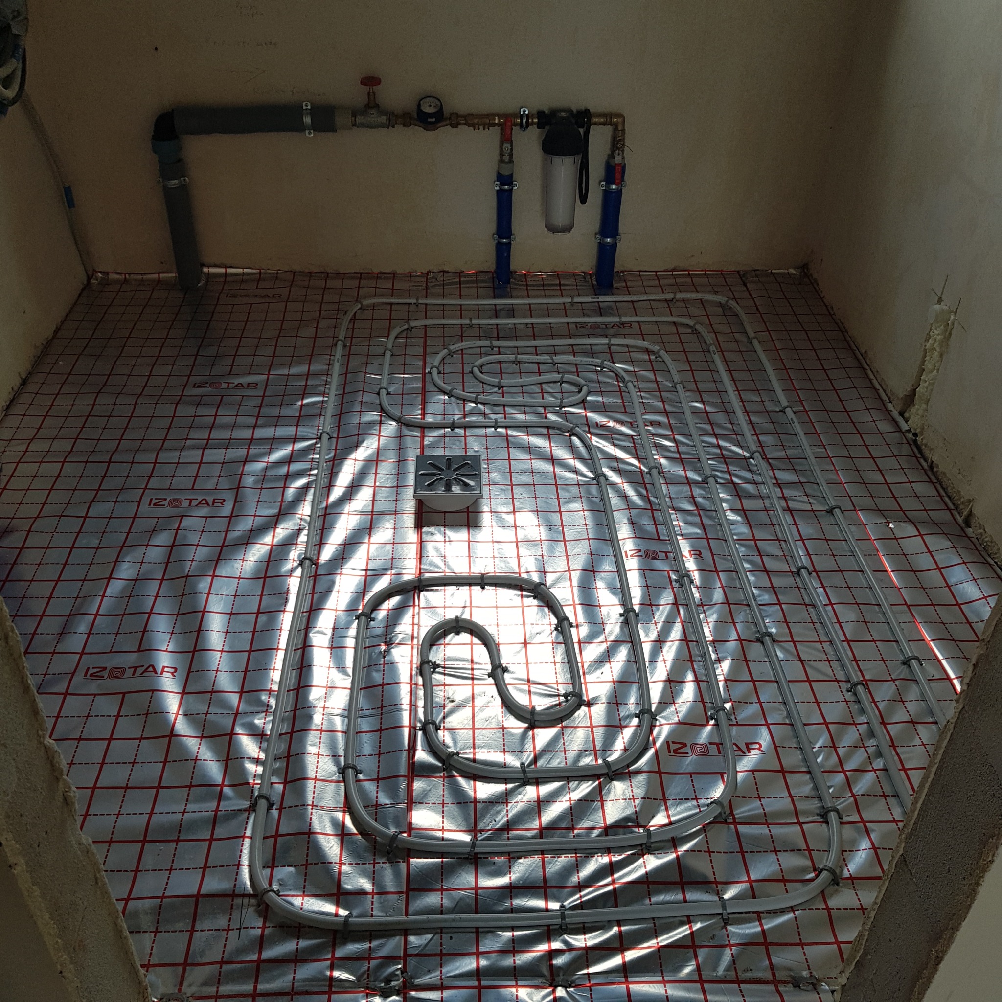 Ułożona instalacja ogrzewania podłogowego na folii izolacyjnej z widocznym kolektorem i filtrem wody. Szare rury tworzą pętle na srebrnej macie z czerwoną kratką.