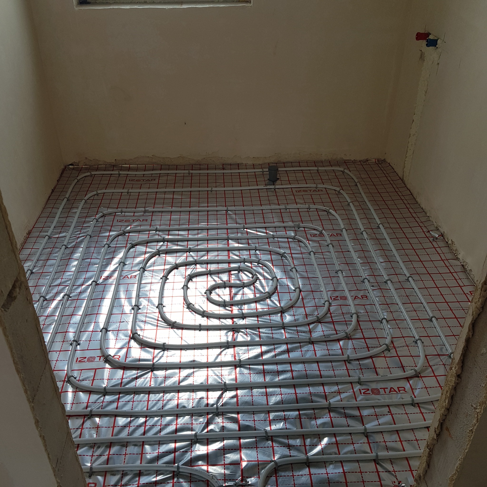 Ułożona instalacja ogrzewania podłogowego na folii izolacyjnej IZOSTAR w surowym pomieszczeniu. Widoczne rury w kształcie spirali i mocowania.