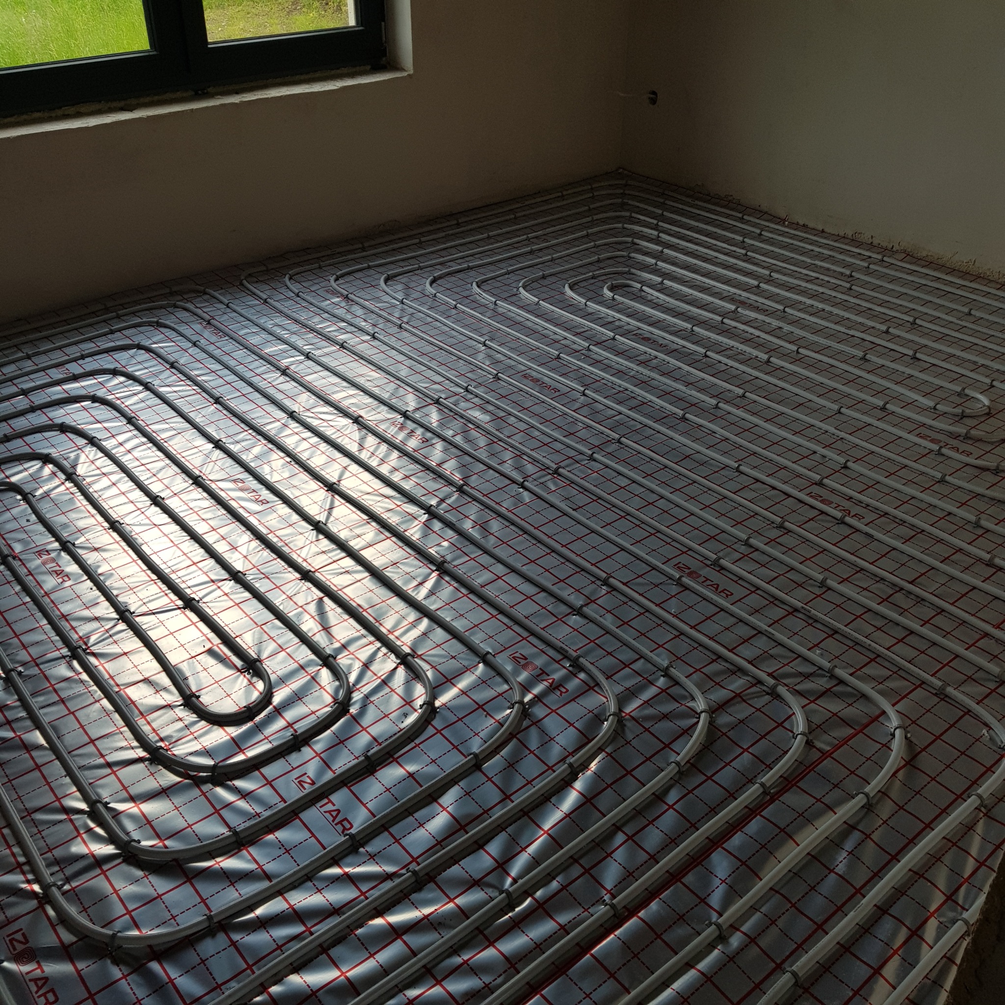 Ułożone rury ogrzewania podłogowego na folii z czerwoną kratką, widoczne okno w tle. Instalacja w jasnym pomieszczeniu przed wylaniem posadzki.