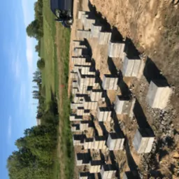 Przygotowane betonowe bloki pod fundamenty, ułożone w rzędach na placu budowy, z widocznym samochodem dostawczym w tle.