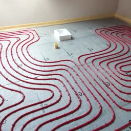 G-HEAT INSTALACJE HYDRAULICZNE - Czerwone rurki ogrzewania podłogowego ułożone na izolacji z folią aluminiową, biały styropianowy element i żółta rolka taśmy, widoczne rury instalacji na ścianie.