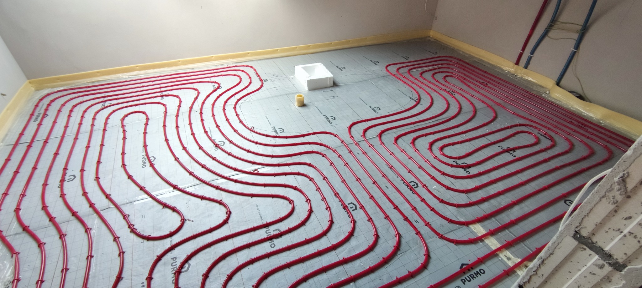 Czerwone rurki ogrzewania podłogowego ułożone na izolacji z folią aluminiową, biały styropianowy element i żółta rolka taśmy, widoczne rury instalacji na ścianie.