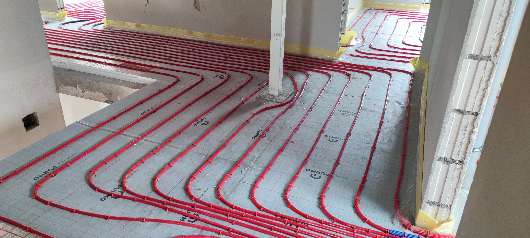 Czerwone rury grzewcze ułożone na szarych płytach izolacyjnych z nadrukowanym logo PURMO, widok z góry na etapie instalacji w surowym stanie budynku.