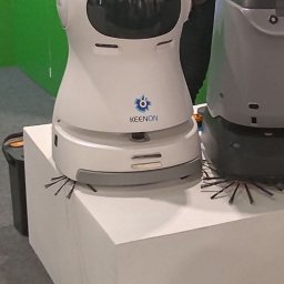 Tetiana Ivankova Global Clean Kraków - Biały robot sprzątający Keenon z kocimi uszami na białym postumencie, obok szarego robota. Widoczne szczotki do zamiatania podłogi. Prezentacja na targach.