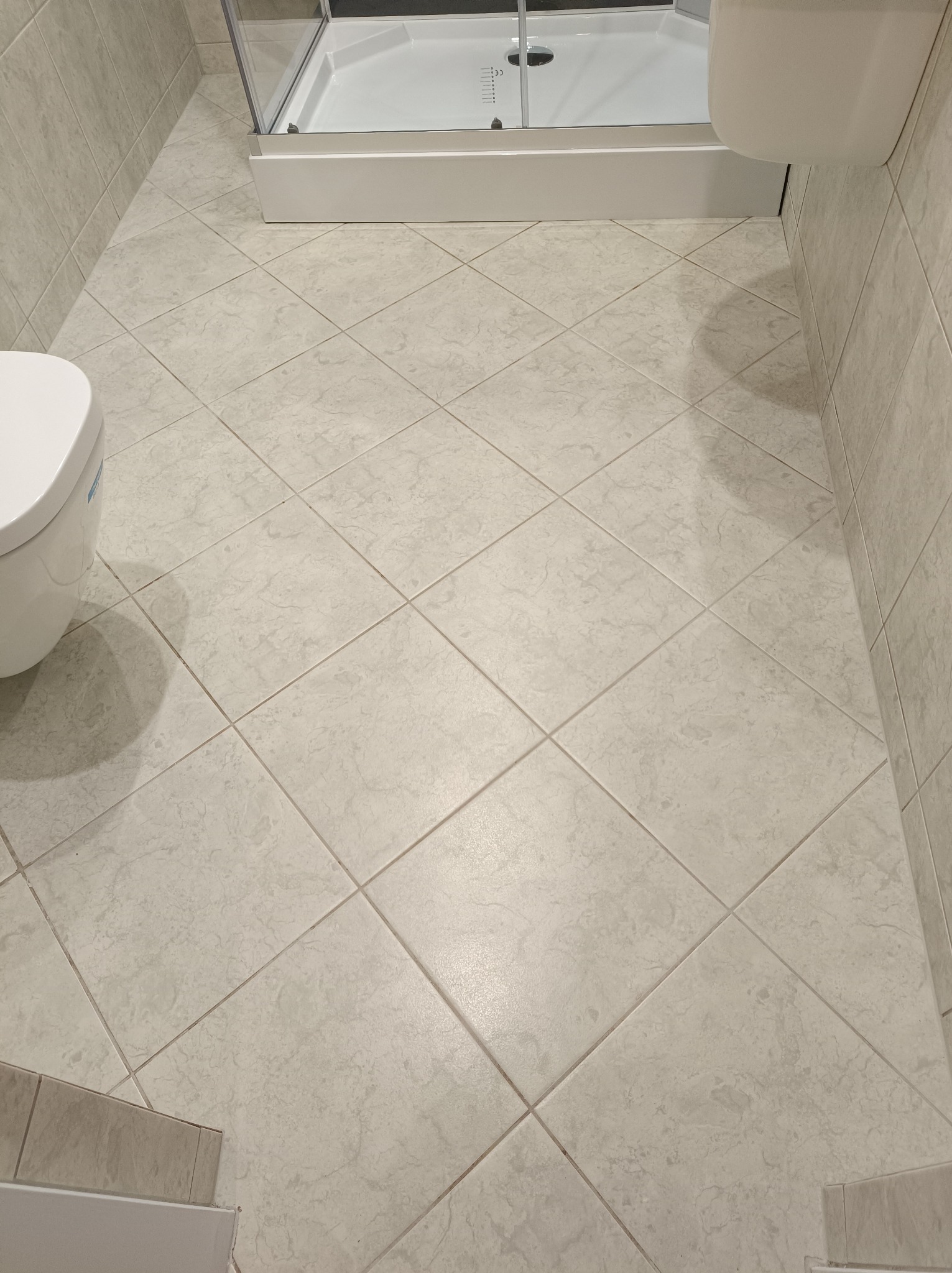 Jasne, kwadratowe płytki podłogowe w łazience, widoczny fragment toalety i kabiny prysznicowej.