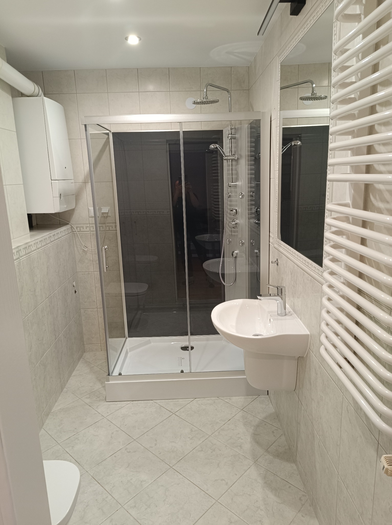 Nowoczesna łazienka z kabiną prysznicową typu walk-in, umywalką na postumencie i grzejnikiem drabinkowym, wykończona jasnymi płytkami.