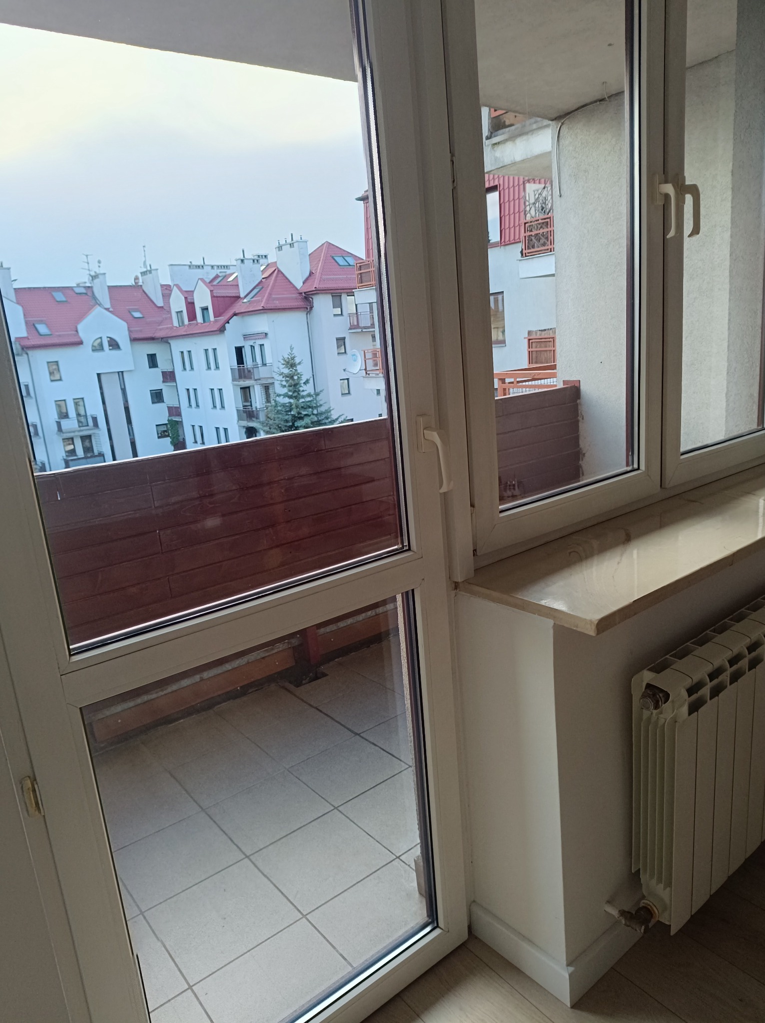 Otwarty balkon z widokiem na budynki mieszkalne z czerwonymi dachami, widoczna grzejnik i parapet wewnątrz pomieszczenia.