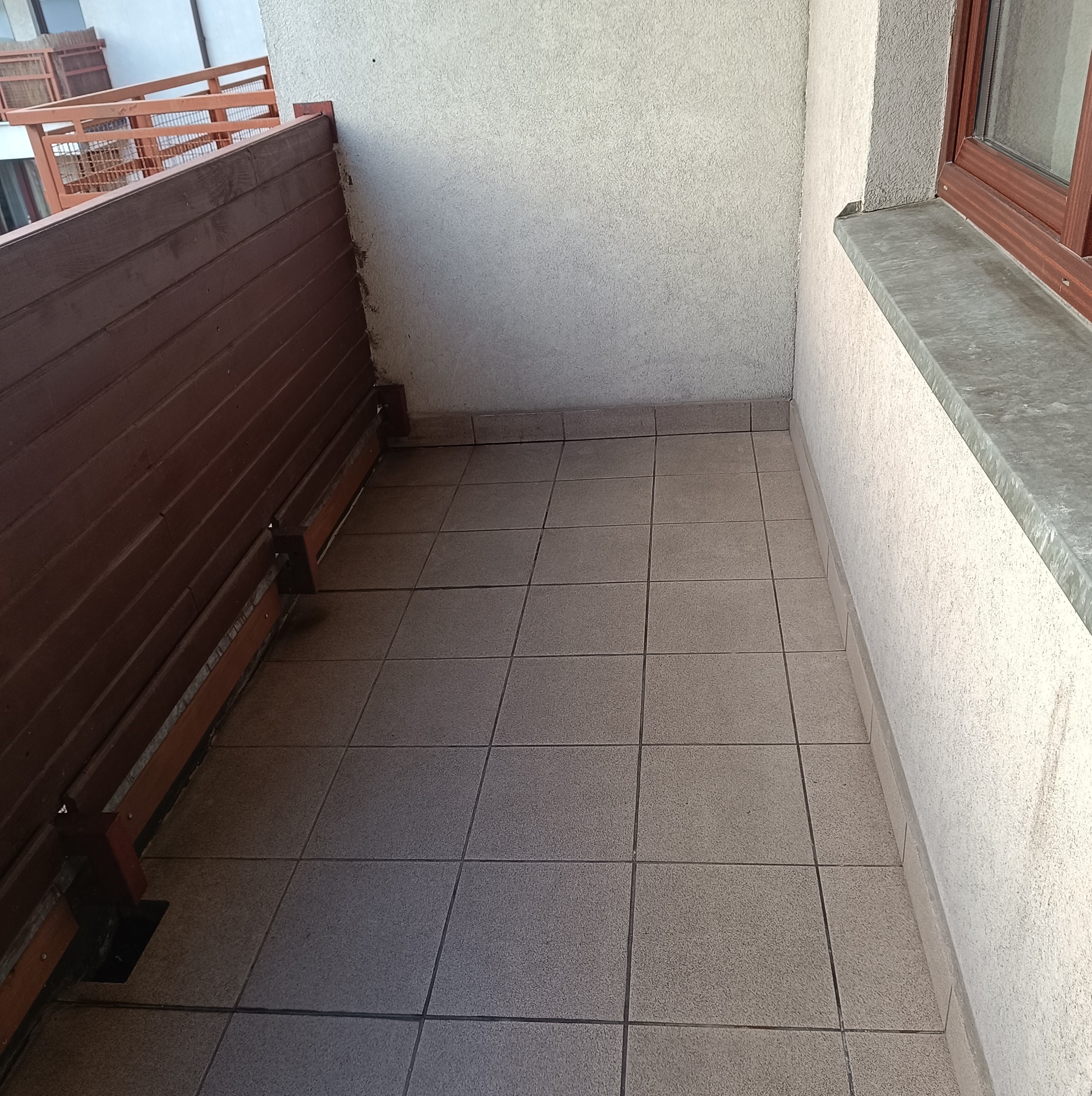 Pusty balkon z brązową drewnianą barierką, szarymi płytkami podłogowymi i oknem po prawej stronie, widok z góry.