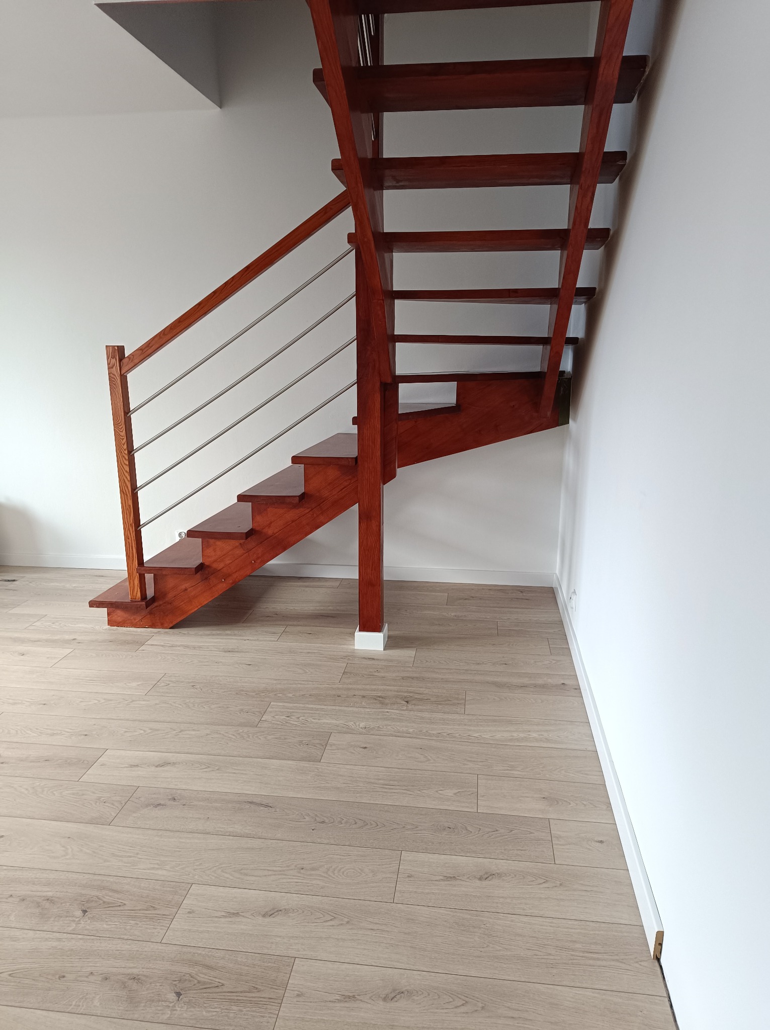 Drewniane schody z metalową balustradą na tle białych ścian i podłogi z paneli imitujących drewno.