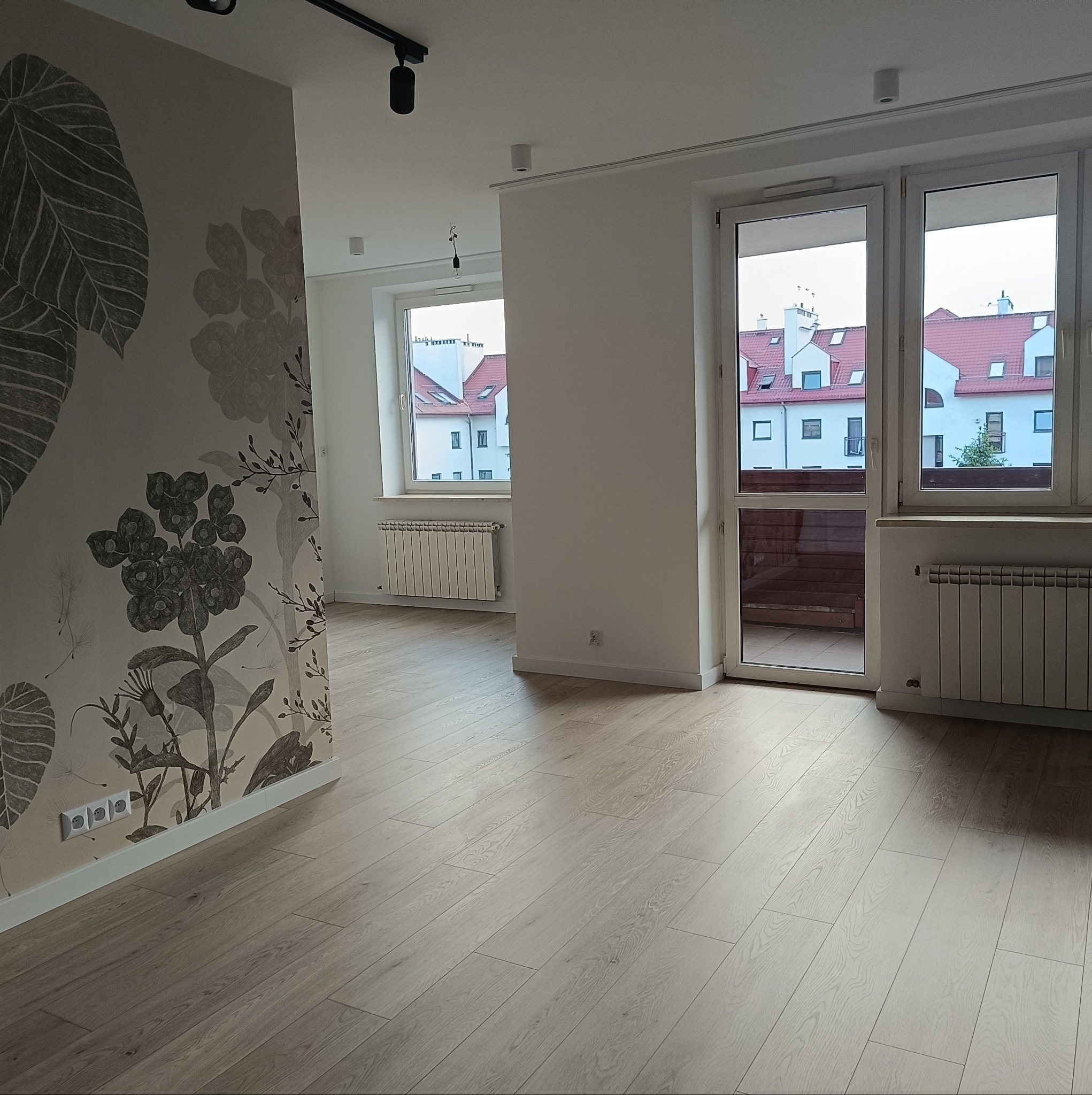 Puste, jasne wnętrze mieszkania z drewnianą podłogą, grzejnikami pod oknami i tapetą w motywy roślinne na ścianie.