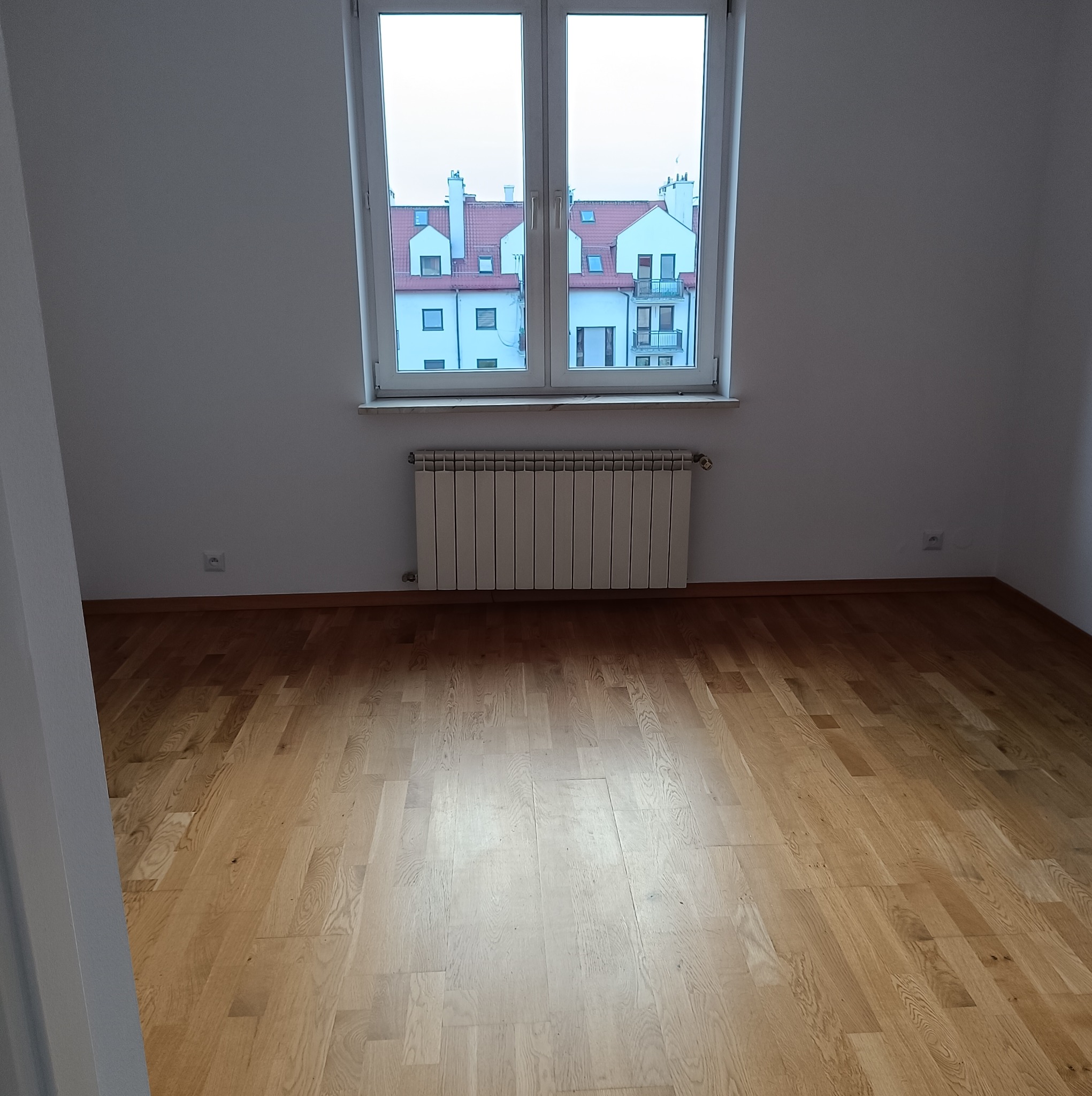 Pusty pokój z parkietem, grzejnikiem pod oknem i widokiem na budynki mieszkalne z czerwonymi dachami.