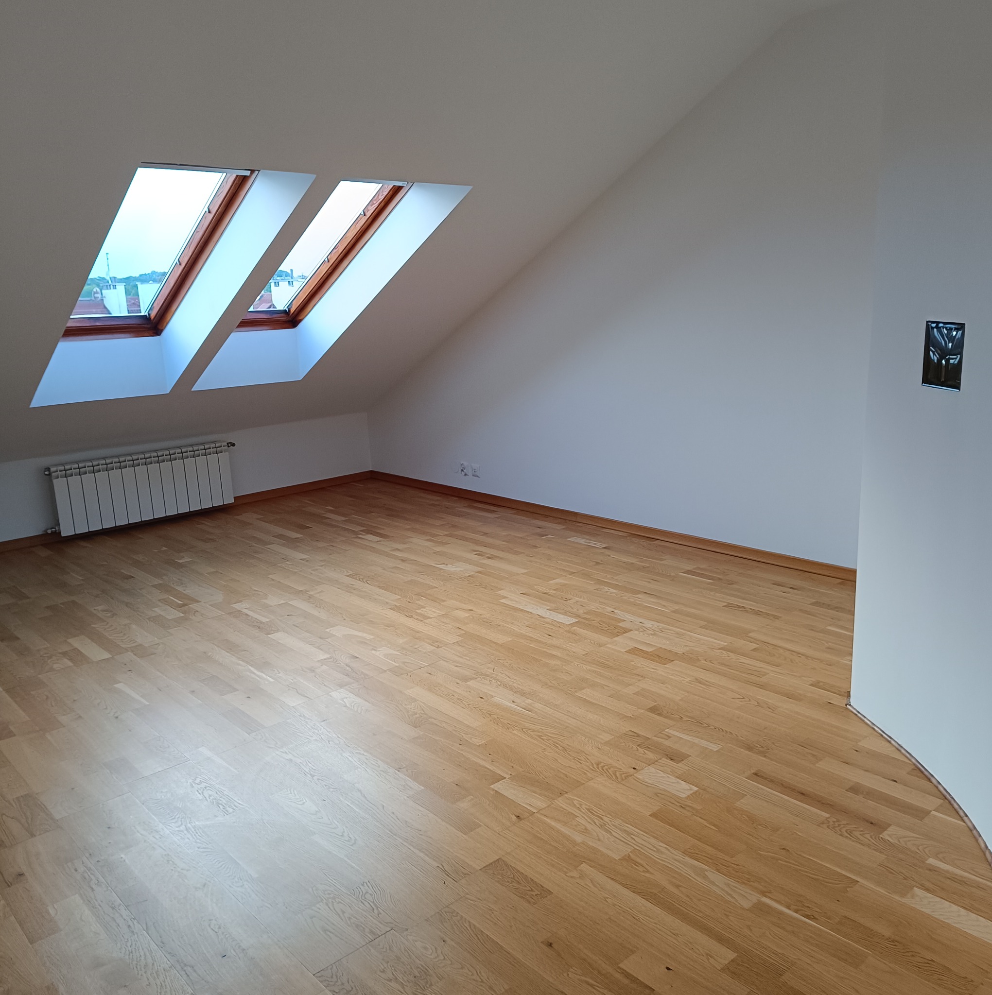 Puste poddasze z trzema oknami dachowymi, grzejnikiem i parkietem ułożonym w jodełkę, przygotowane do umeblowania.