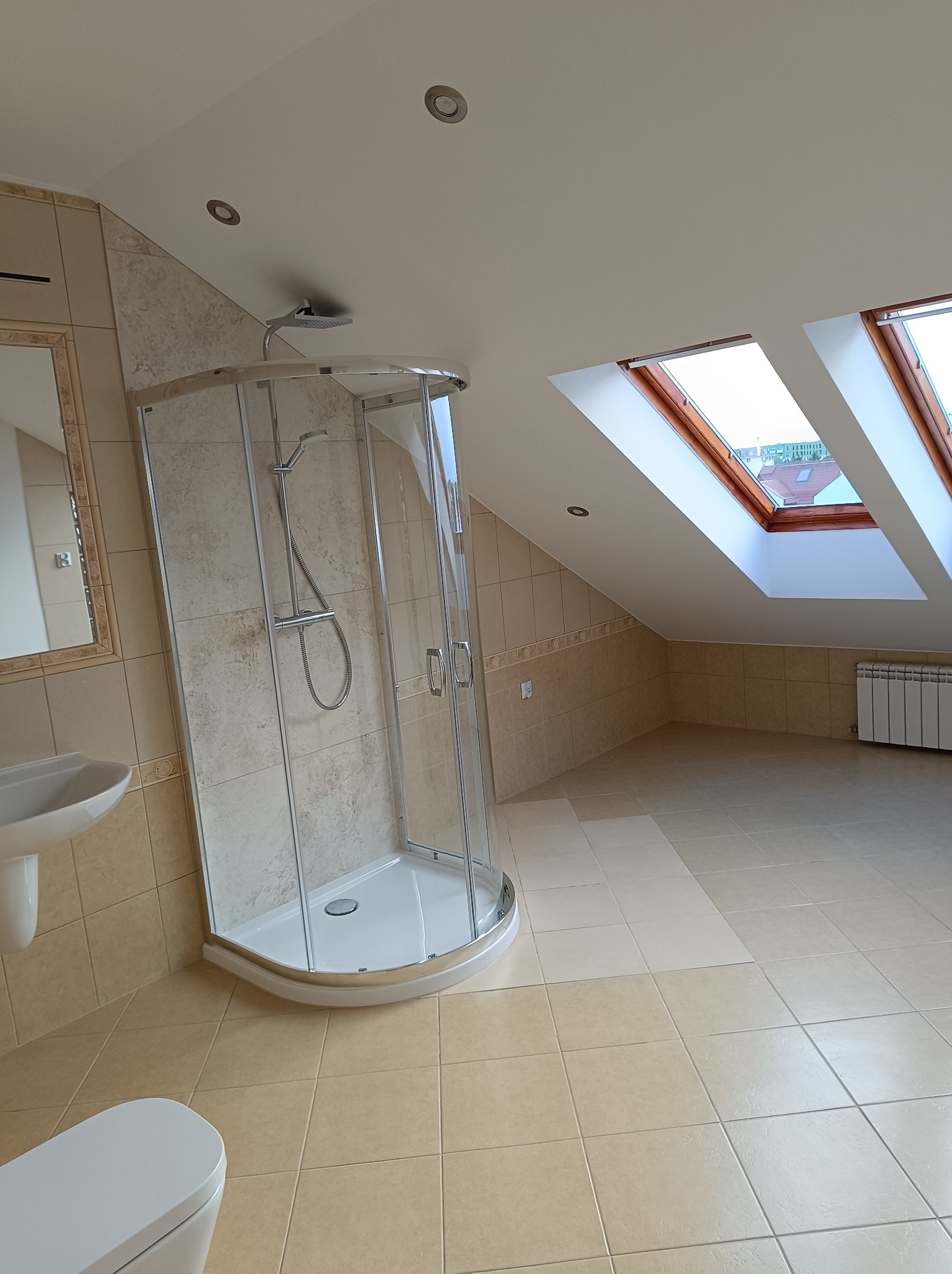 Jasna łazienka na poddaszu z kabiną prysznicową narożną, umywalką na postumencie i dwoma oknami dachowymi w drewnianych ramach.