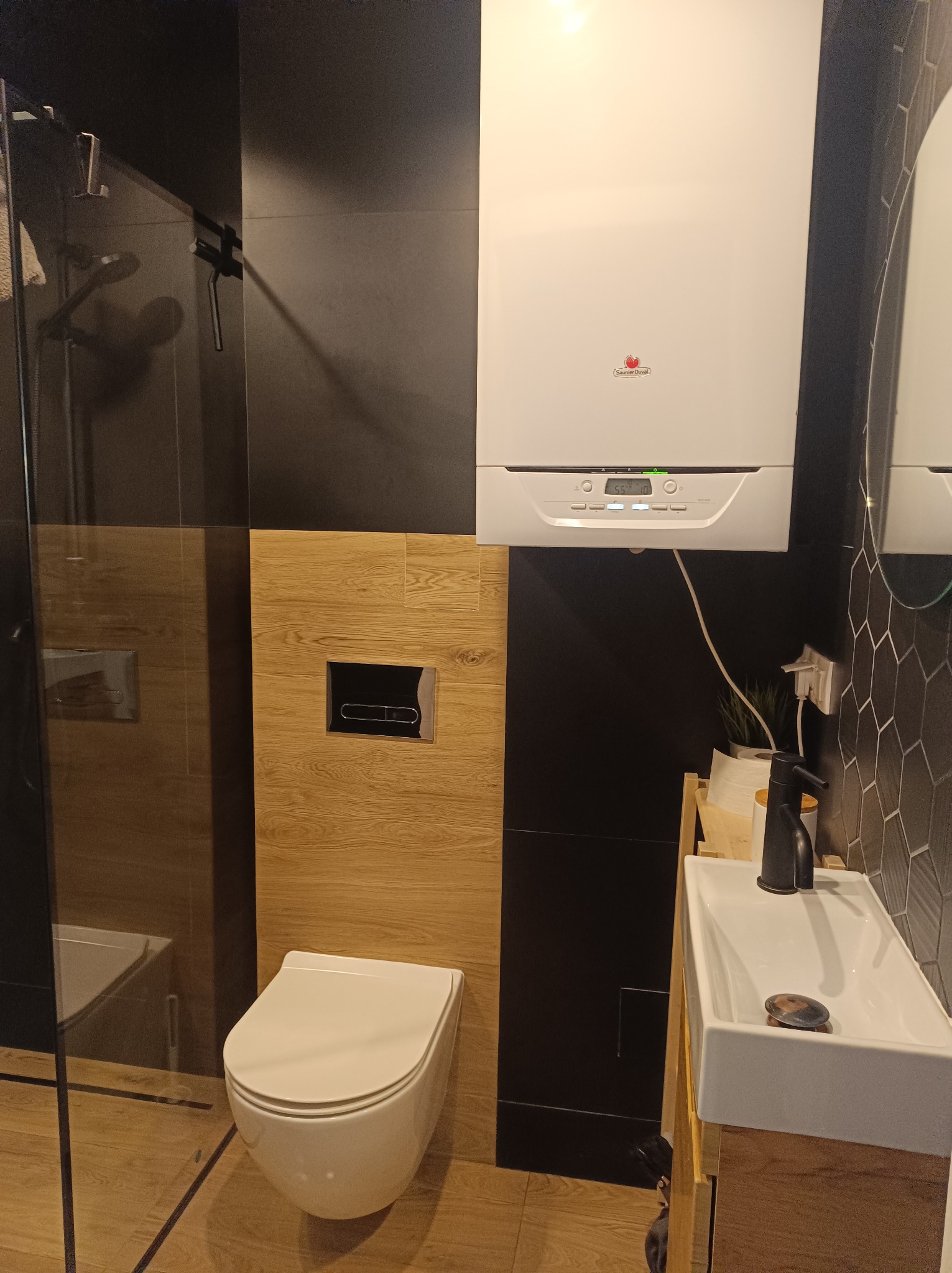 Nowoczesna łazienka z wiszącą toaletą, umywalką na drewnianej szafce i piecem gazowym Saunier Duval na czarnej ścianie z heksagonalnymi płytkami.
