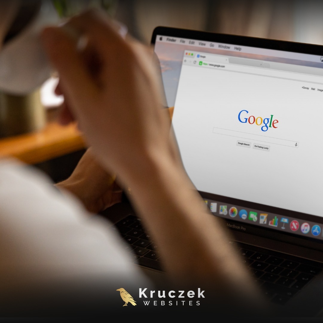 Osoba pracuje na laptopie MacBook Pro, na ekranie widoczna strona główna wyszukiwarki Google. W tle rozmazany kubek. W dolnej części logo firmy Kruczek Websites.