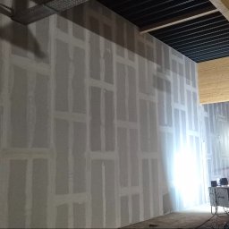 Usługi ogolnobudowlane - Ściana z płyt kartonowo-gipsowych w trakcie szpachlowania, widoczne łączenia. W tle podnośnik i sprzęt budowlany. Oświetlenie robocze.