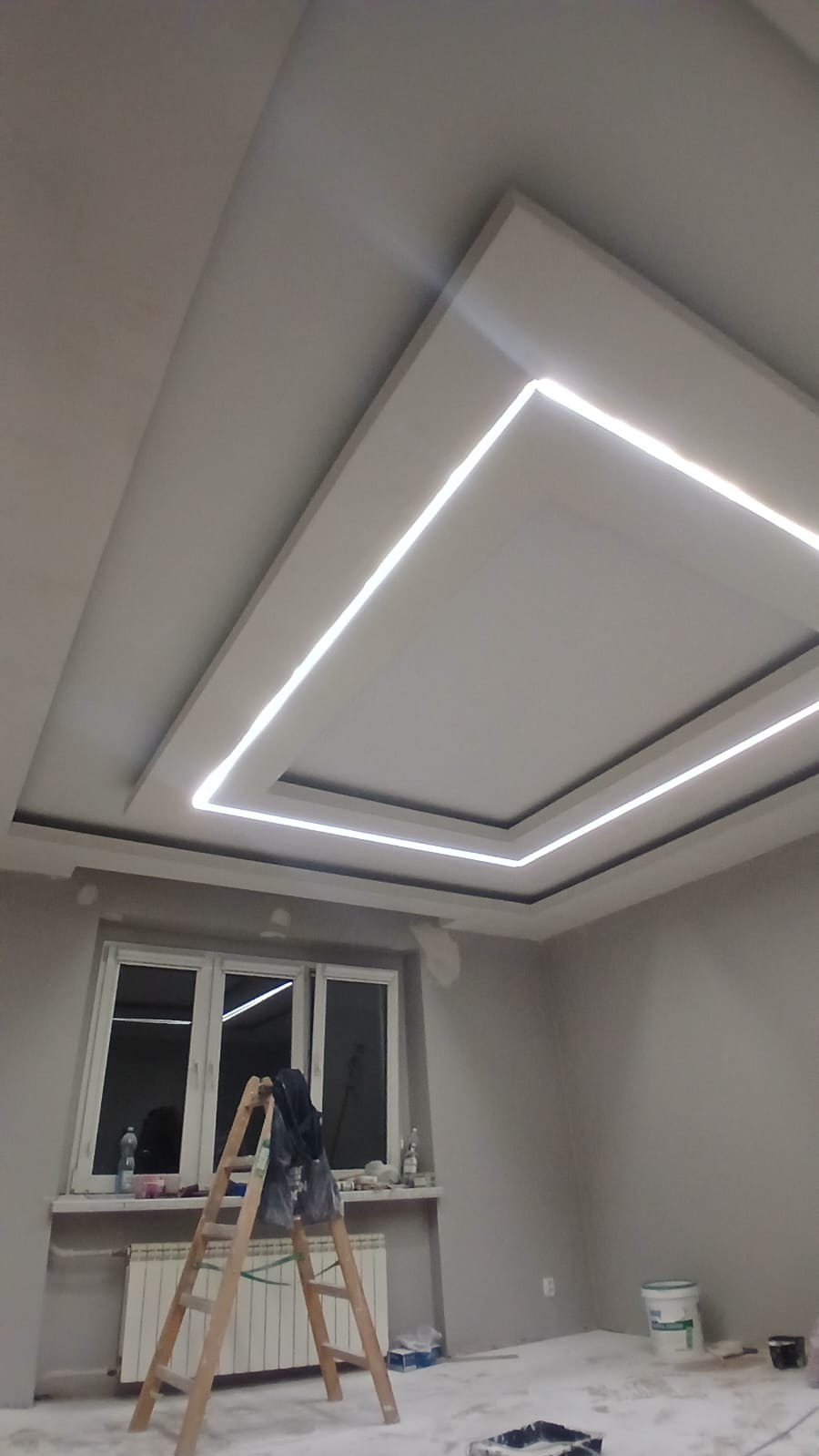 Nowoczesny sufit podwieszany z oświetleniem LED w kształcie prostokąta, drabina malarska w pokoju, szare ściany, okno z parapetem. Widok z dołu.