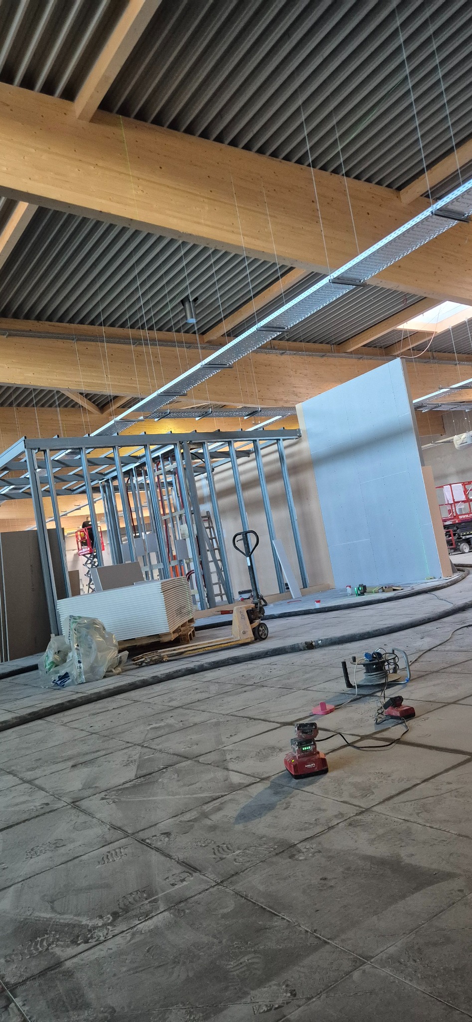 Montaż ścian działowych z metalowych profili i płyt w hali. Widoczny wózek paletowy z płytami, narzędzia i pracownik na podnośniku. Hala z drewnianą konstrukcją dachu.