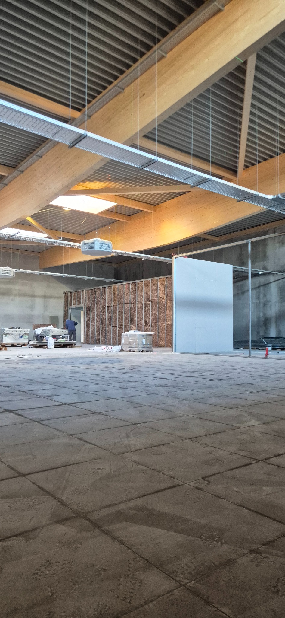 Wnętrze w trakcie prac wykończeniowych: widoczne drewniane belki stropowe, instalacja wentylacyjna, ściana z izolacją i fragment ściany z płytą. W tle majster.